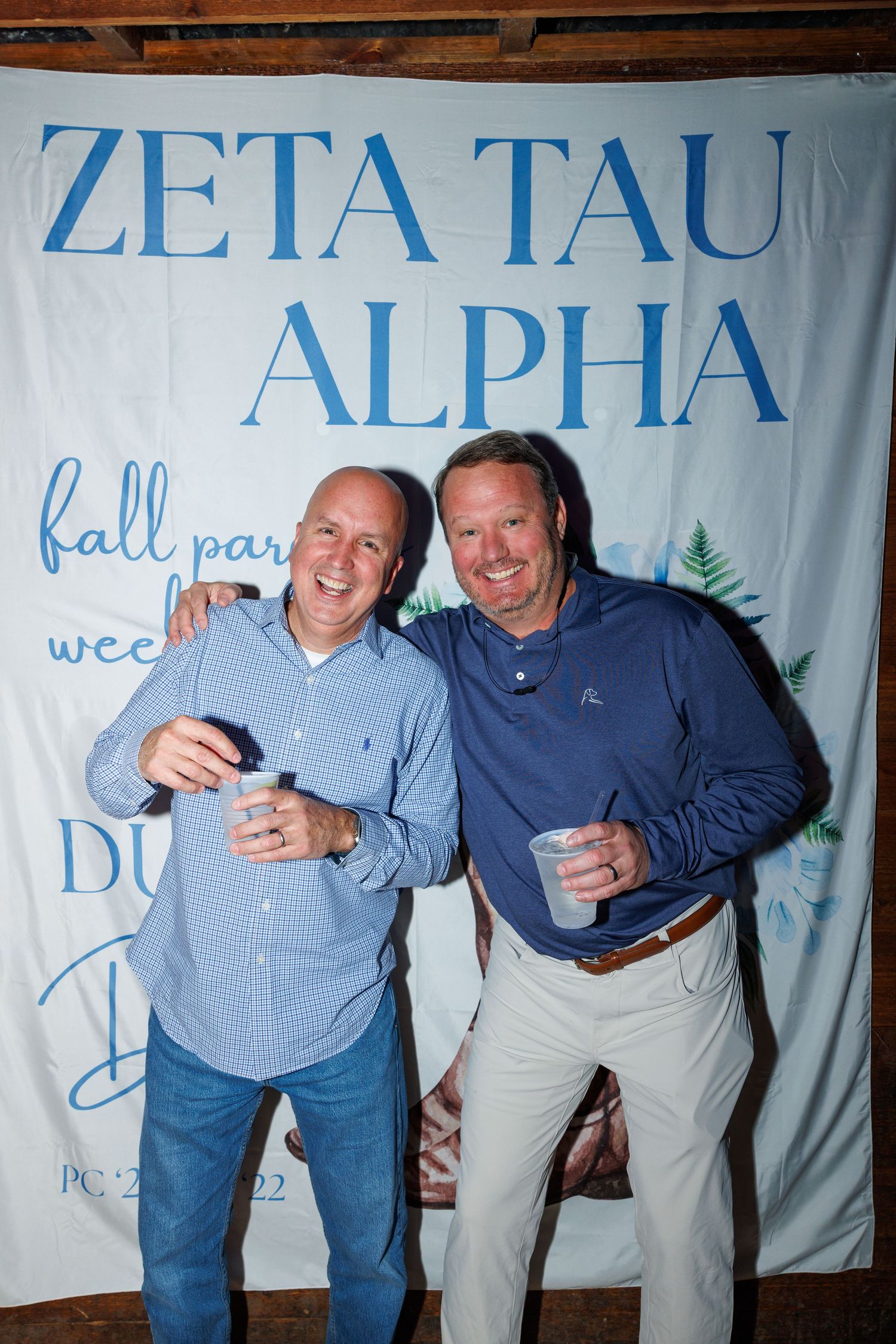 Two men smiling and holding drinks at a Zeta Tau Alpha fall party event backdrop.