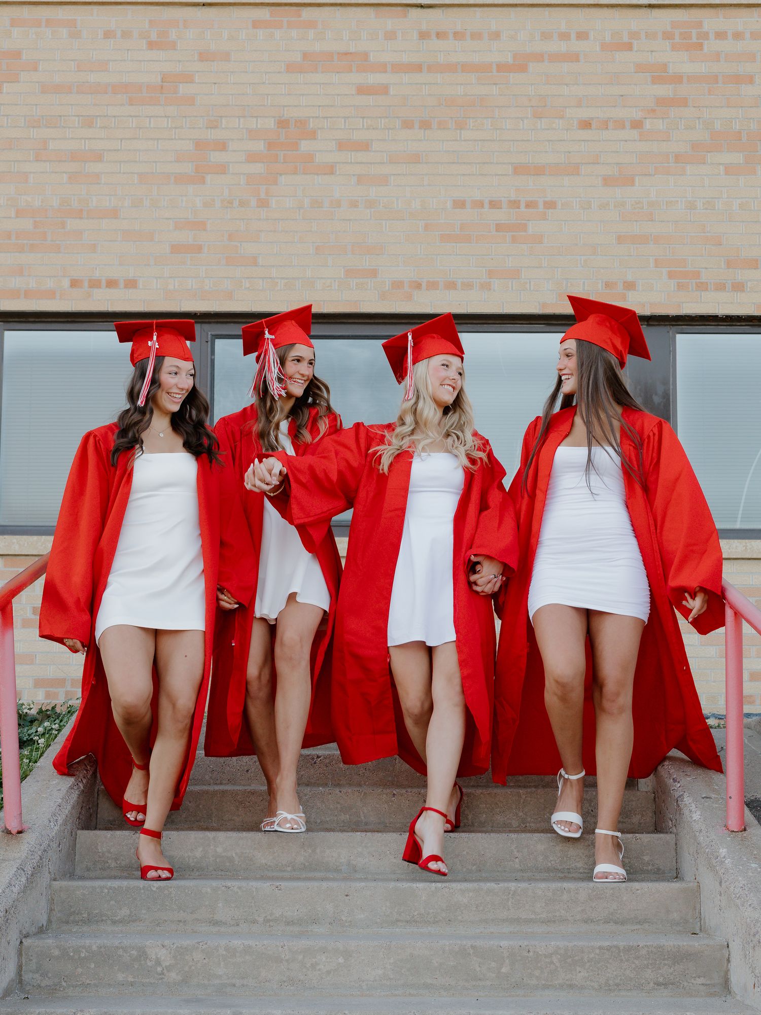 Four girls in red graduation caps and gowns over white dresses walk down steps, smiling and holding hands.
