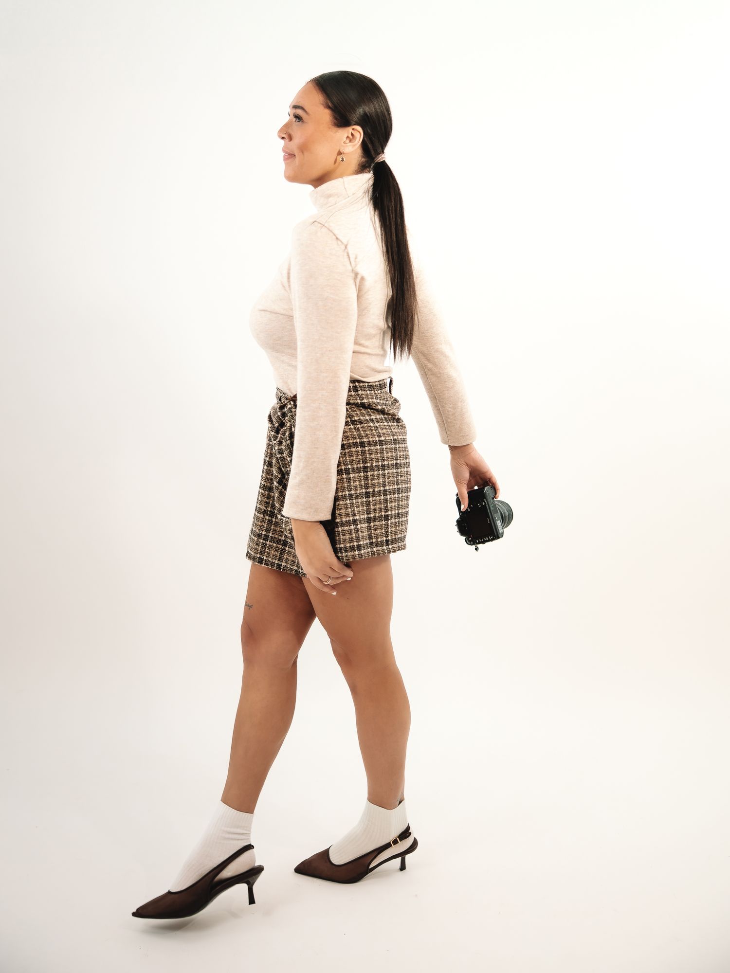 Model in cream turtleneck and plaid shorts poses with black handbag.