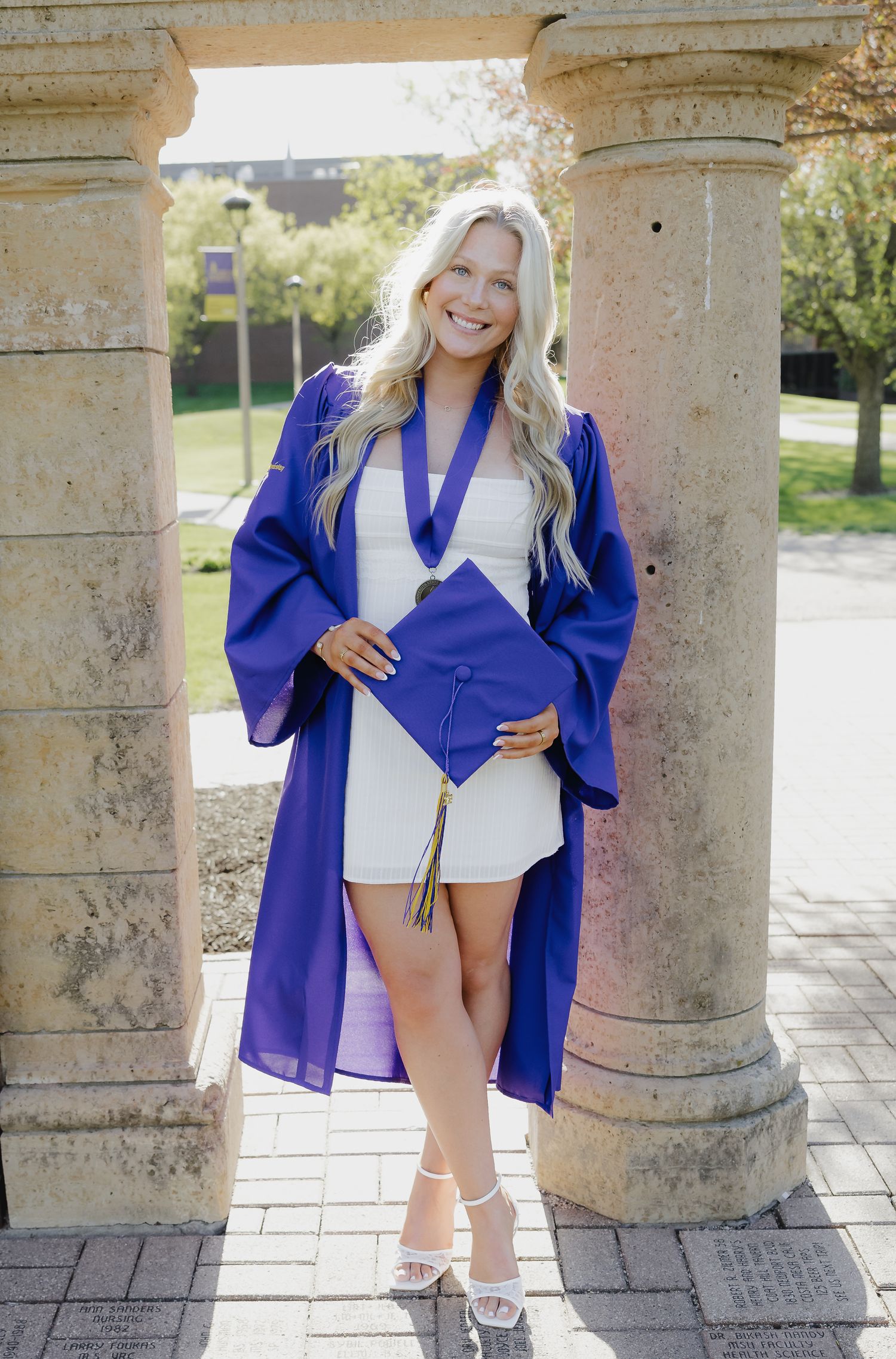 Smiling blonde graduate in purple cap and gown holding mortarboard between stone columns outdoors.