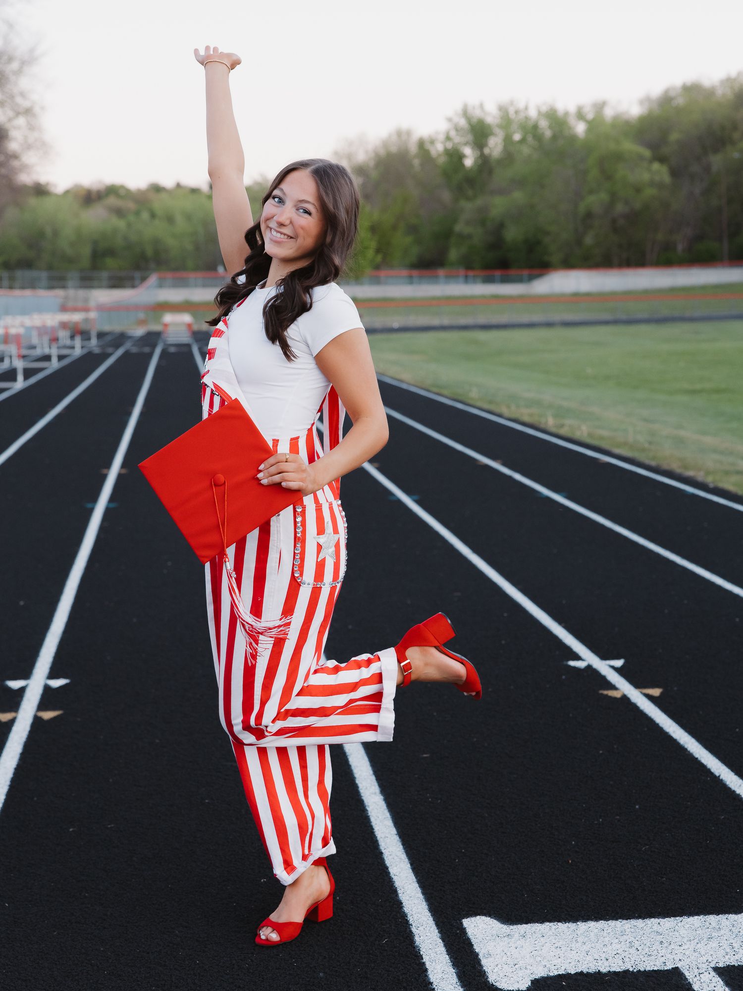 A smiling graduate in red and white striped overalls holds a red cap on a track, celebrating with one arm raised.