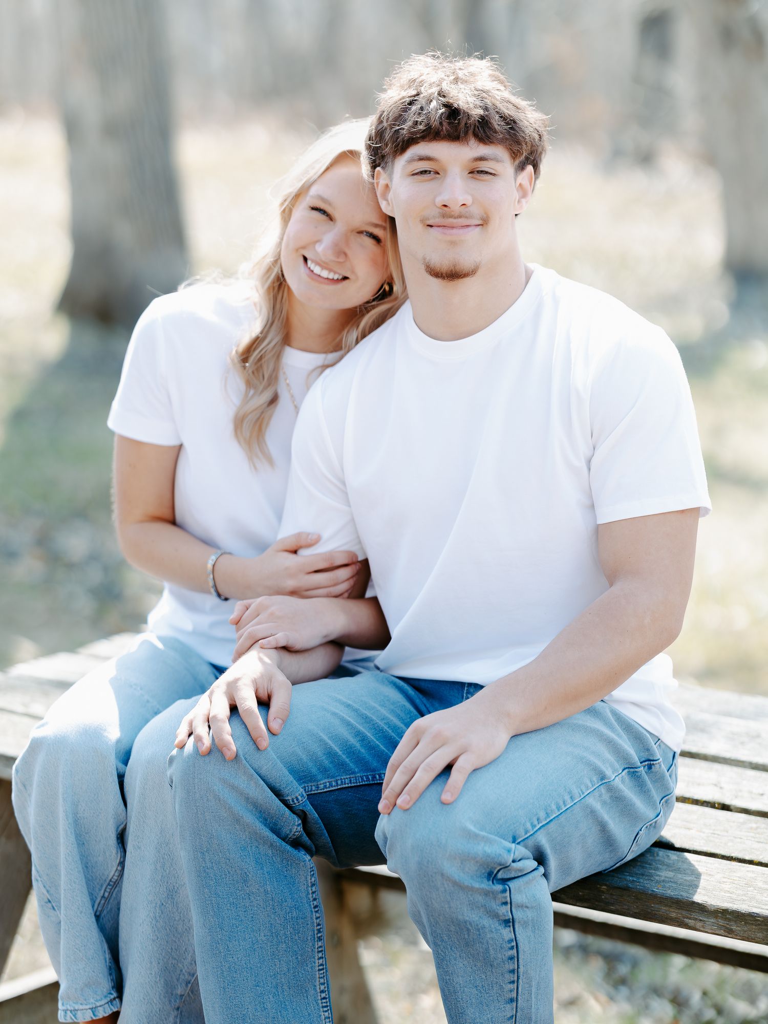 A smiling couple in matching white tees and jeans sit closely on a bench outdoors in a bright, wooded setting.