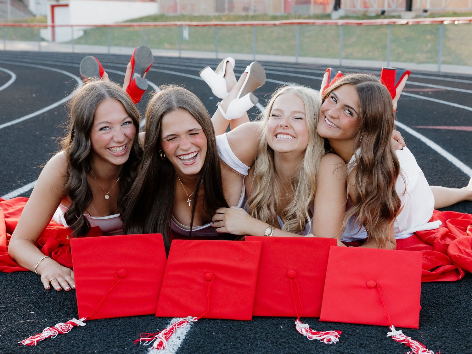 Four smiling young women in red graduation caps lying on a track field celebrating their graduation day.