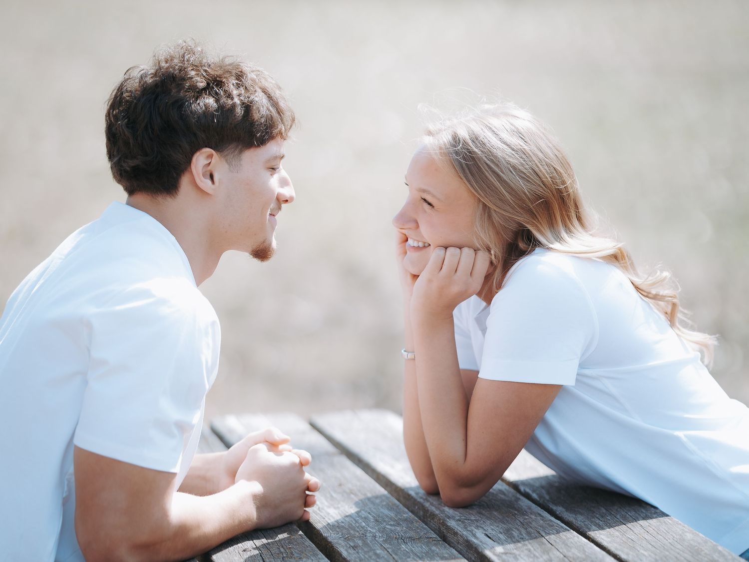 Young couple in white t-shirts smiling at each other while sitting at an outdoor wooden table.