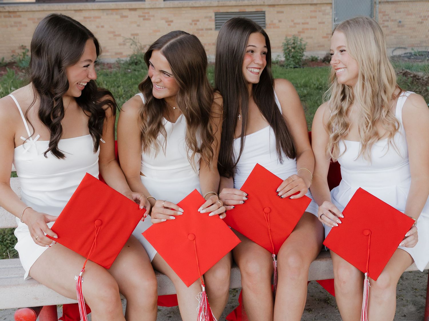 Four young women in white dresses holding red graduation caps sit together on a bench, celebrating their graduation day.