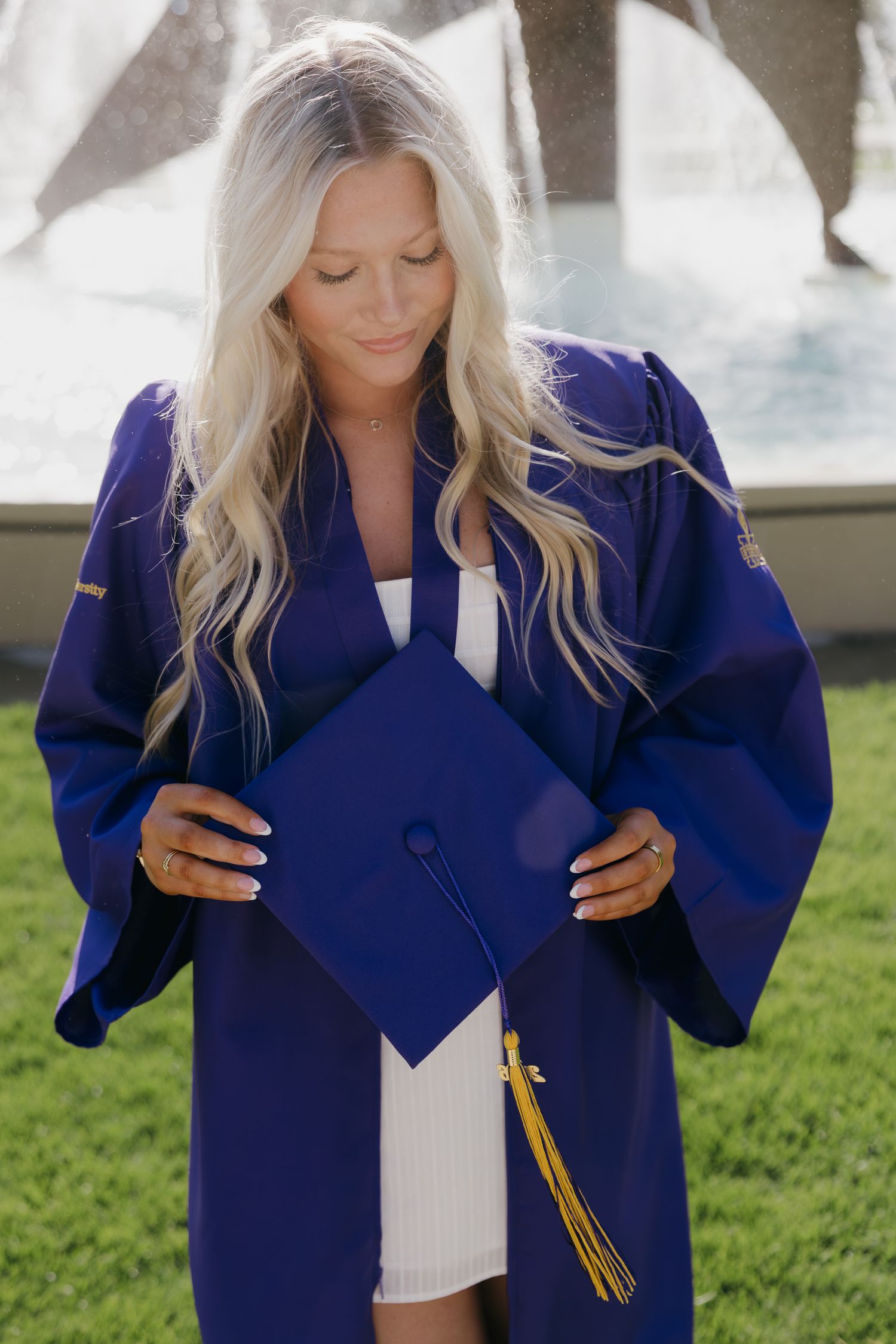 Blonde woman in blue graduation gown holding mortarboard cap with gold tassel outdoors.