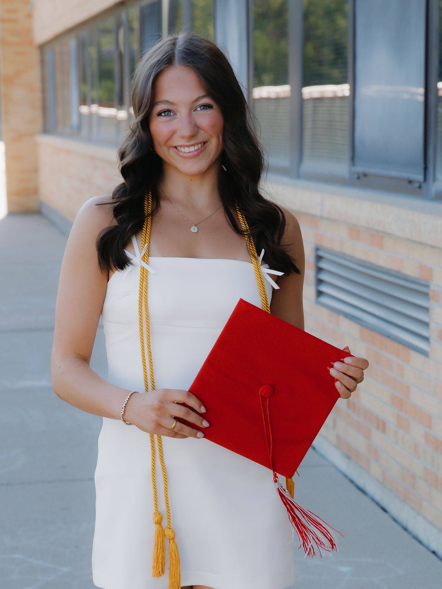Smiling graduate in white dress holds red cap with gold honor cords outdoors near brick building.