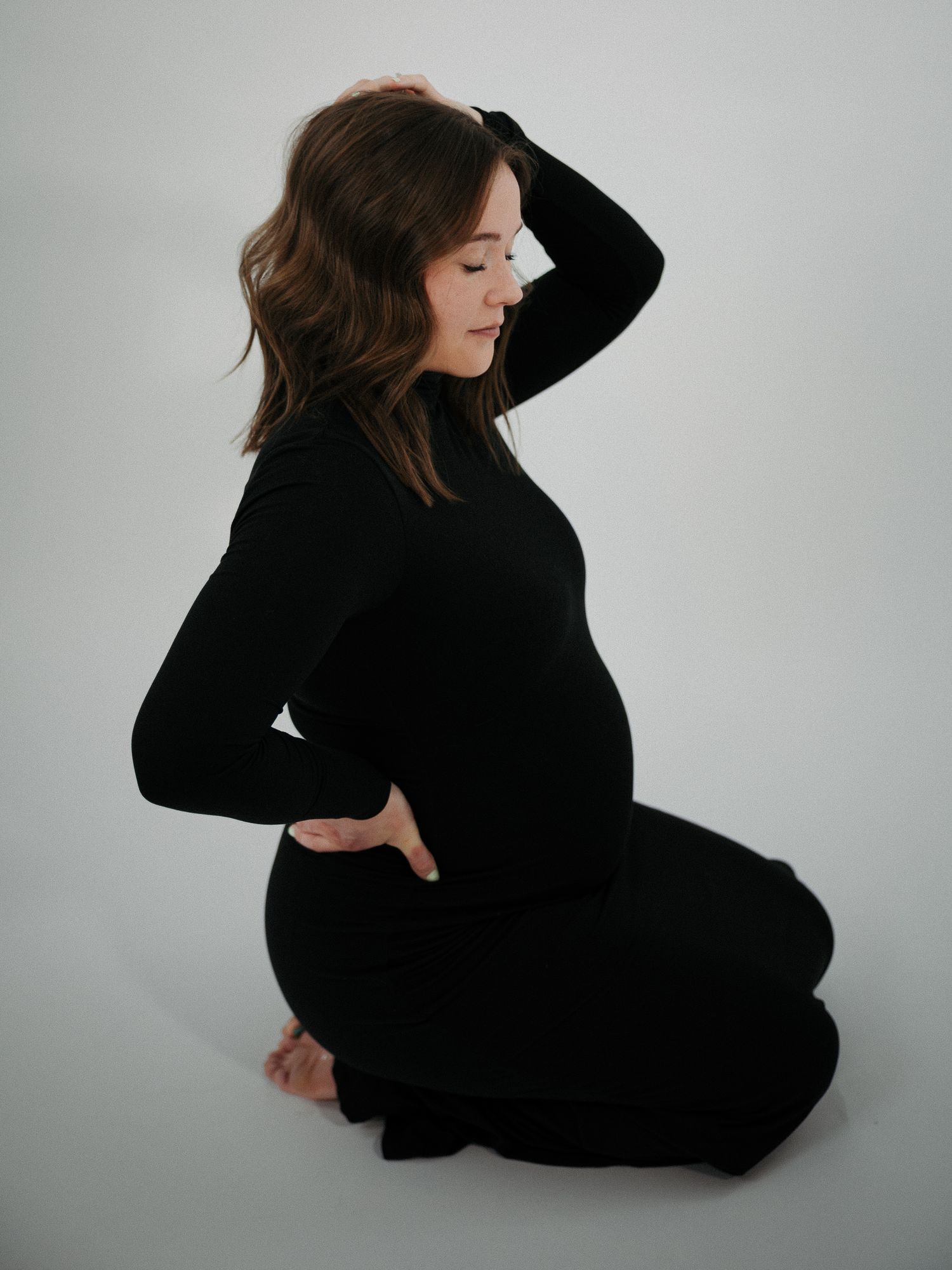 Pregnant woman in black outfit sitting gracefully on floor, hand on back, showcasing baby bump in minimalist studio.