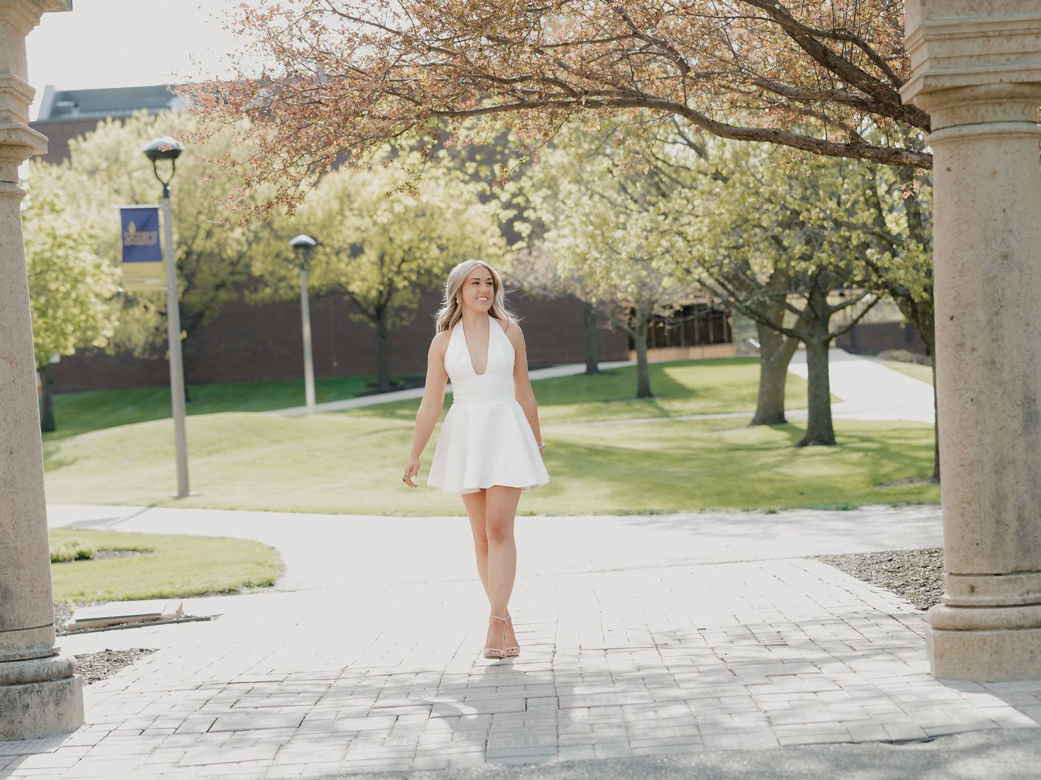 Young woman in white halter dress walking on a sunny campus pathway surrounded by blooming spring trees.