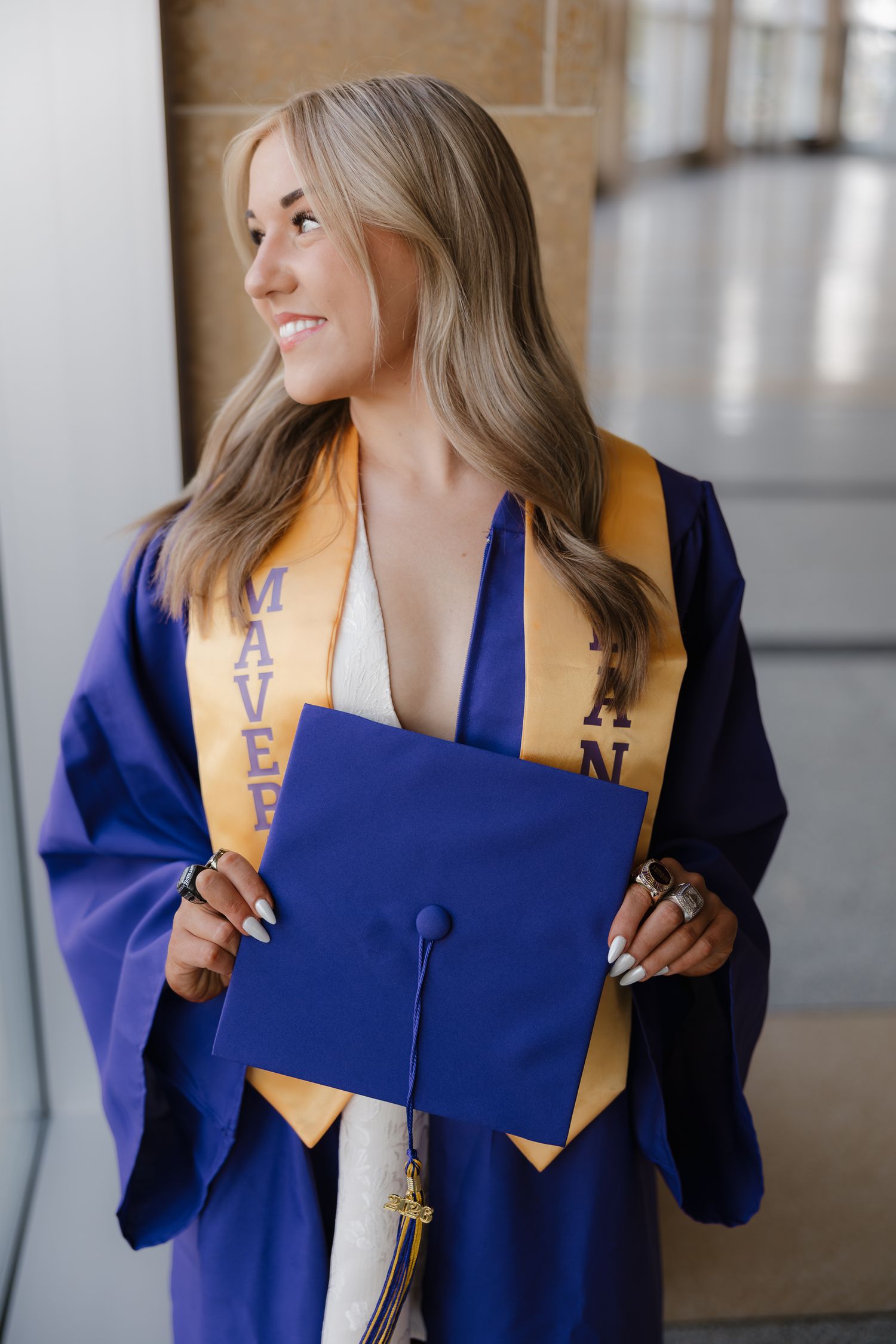 Smiling female graduate in blue cap and gown holding mortarboard, wearing gold Mavericks stole.