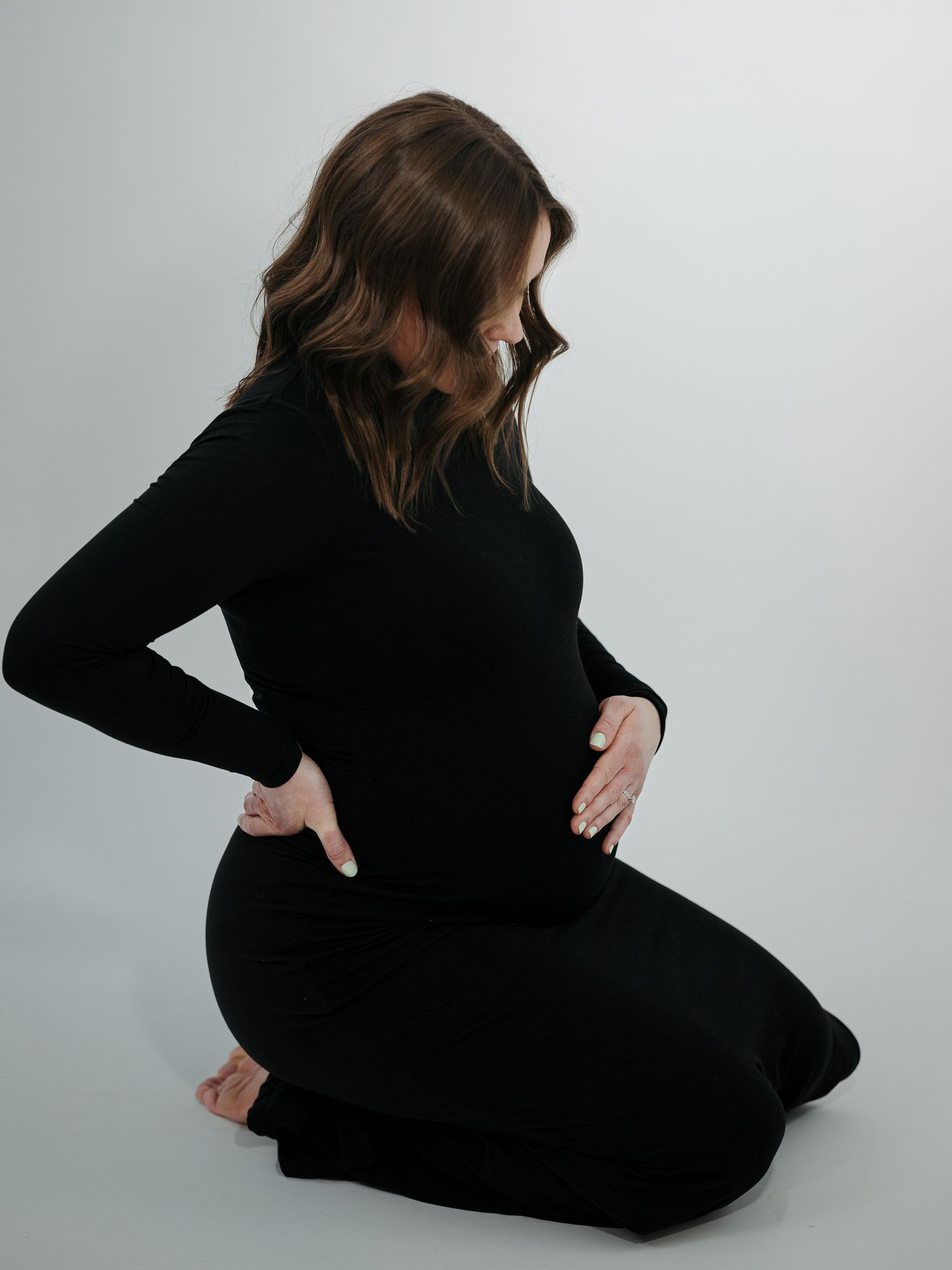 Pregnant woman in black dress kneeling, cradling baby bump with both hands against light gray background.