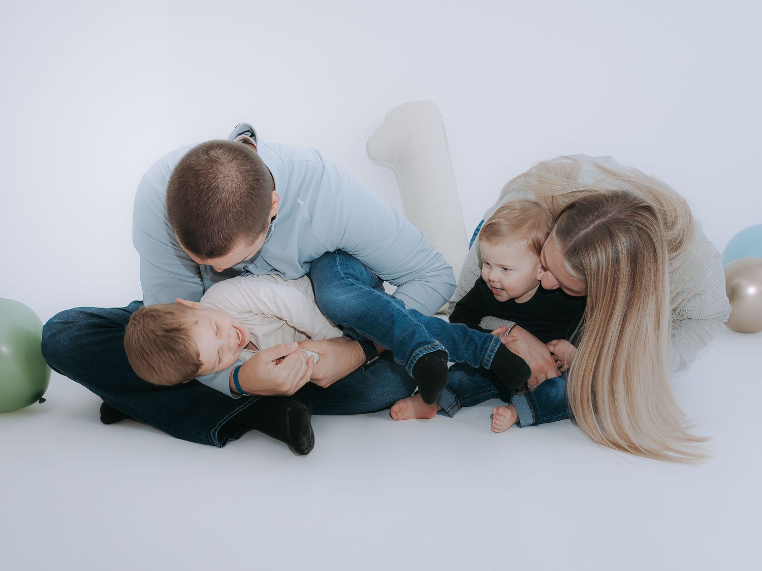 A loving family of four sits on a white floor, parents kissing their two young children surrounded by pastel balloons.