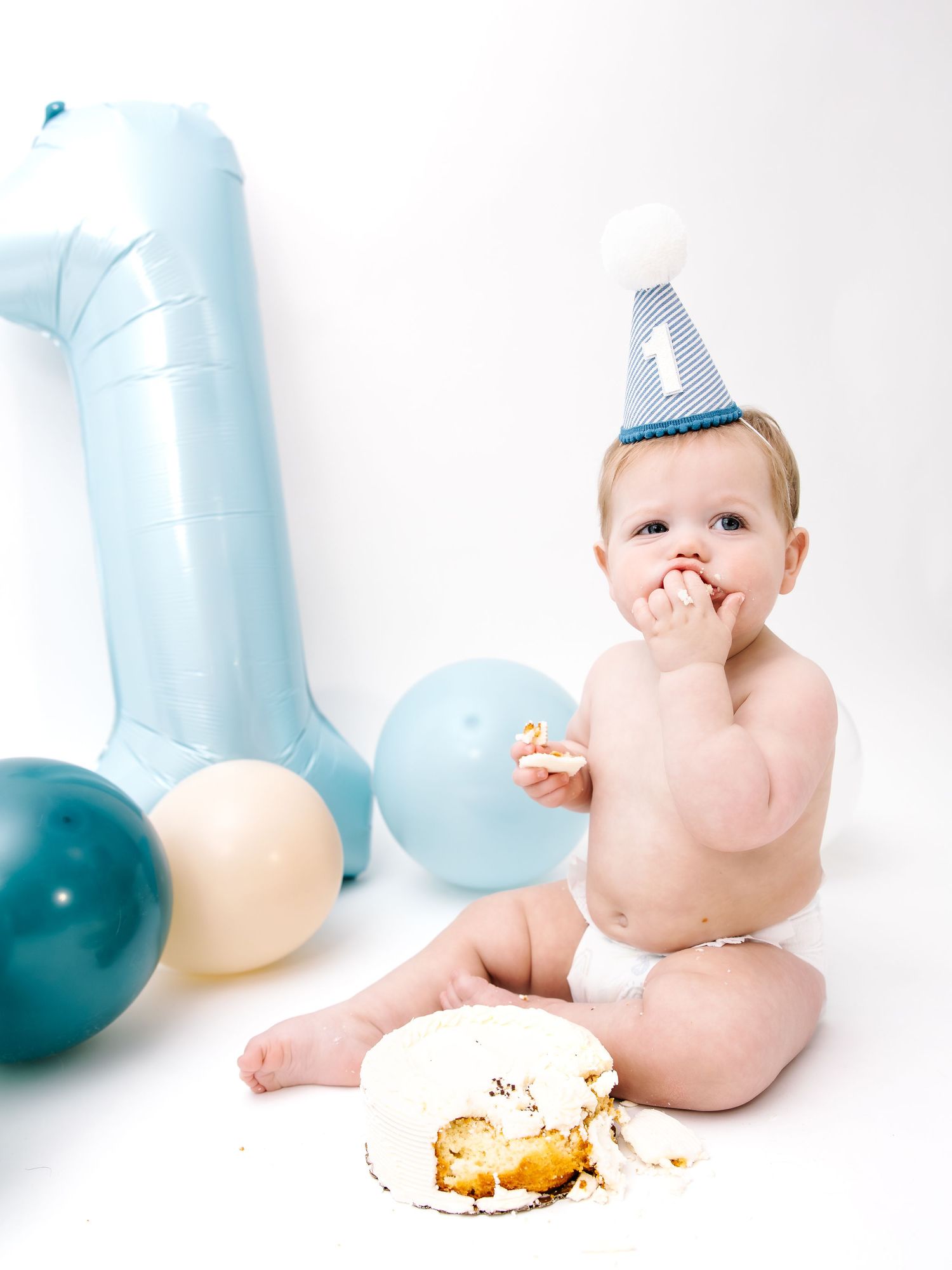 Baby in diaper and blue party hat smashes first birthday cake surrounded by blue balloons and number one balloon.