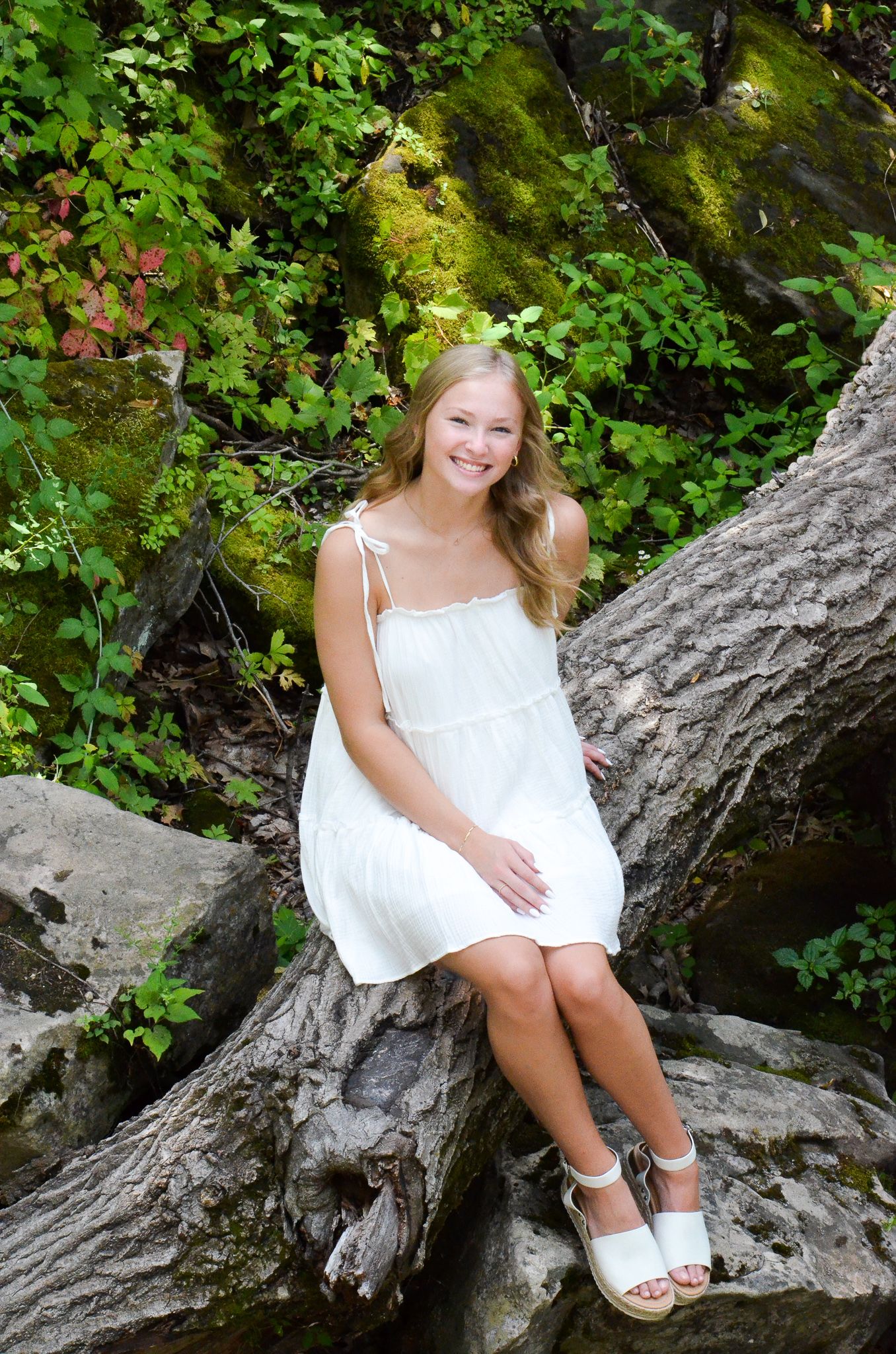 Young person in white dress sitting on mossy rocks surrounded by lush greenery.