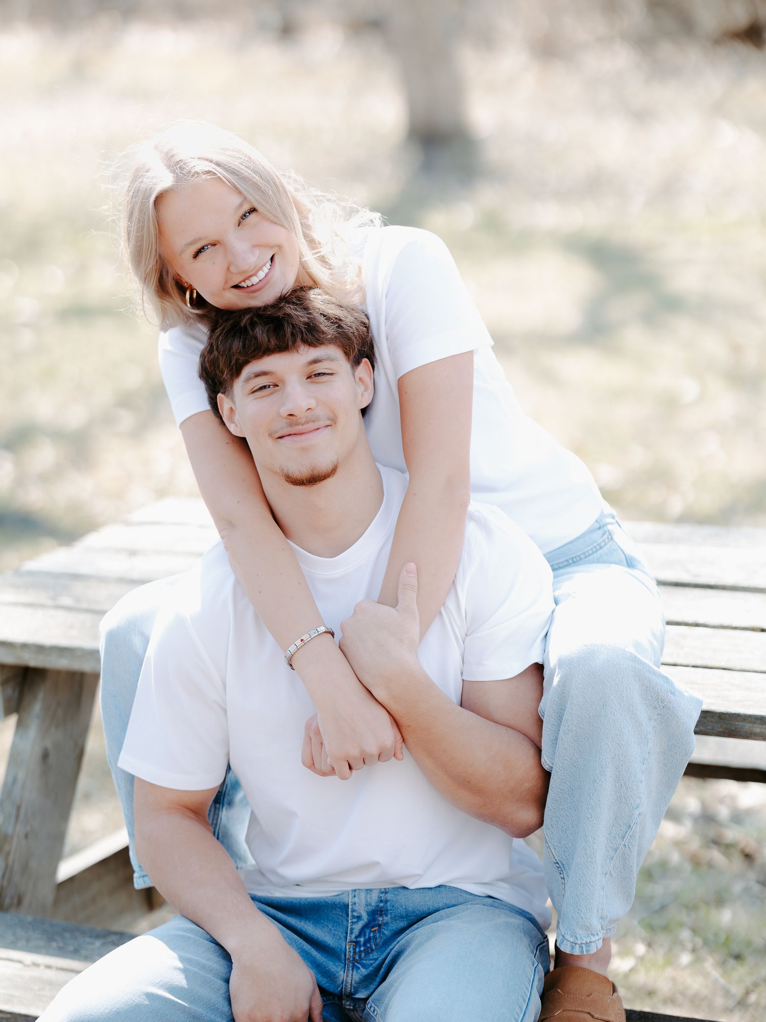A smiling blonde woman hugs a young man from behind as they pose together outdoors on a wooden bench in casual white tees.