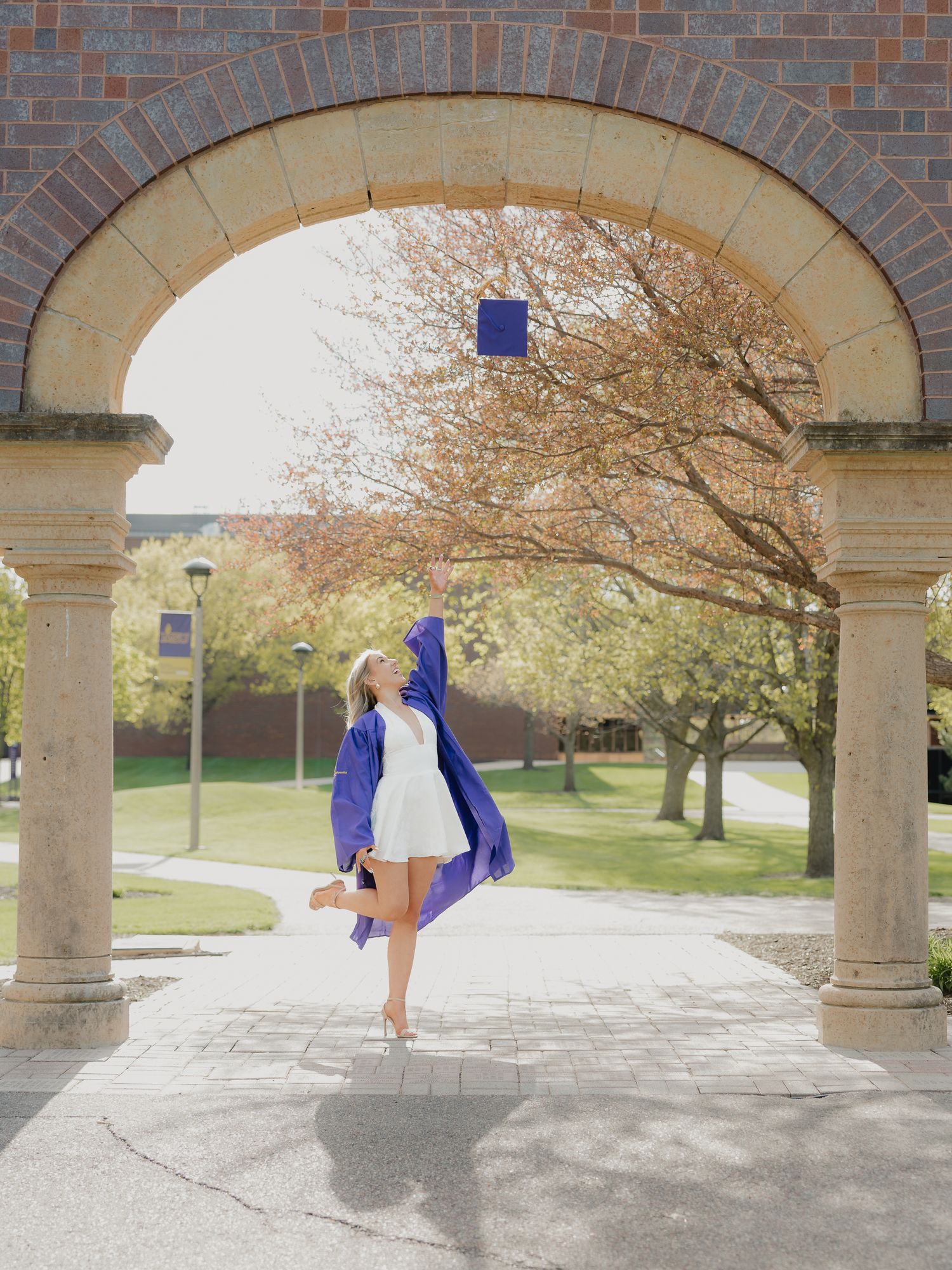 Graduate in blue cap and gown poses joyfully under stone arch on sunny college campus.