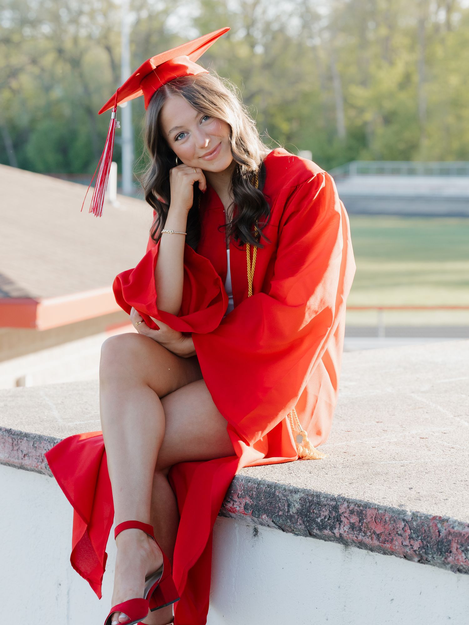 Young woman in red graduation cap and gown sitting outdoors, smiling, wearing red heels at her graduation ceremony.