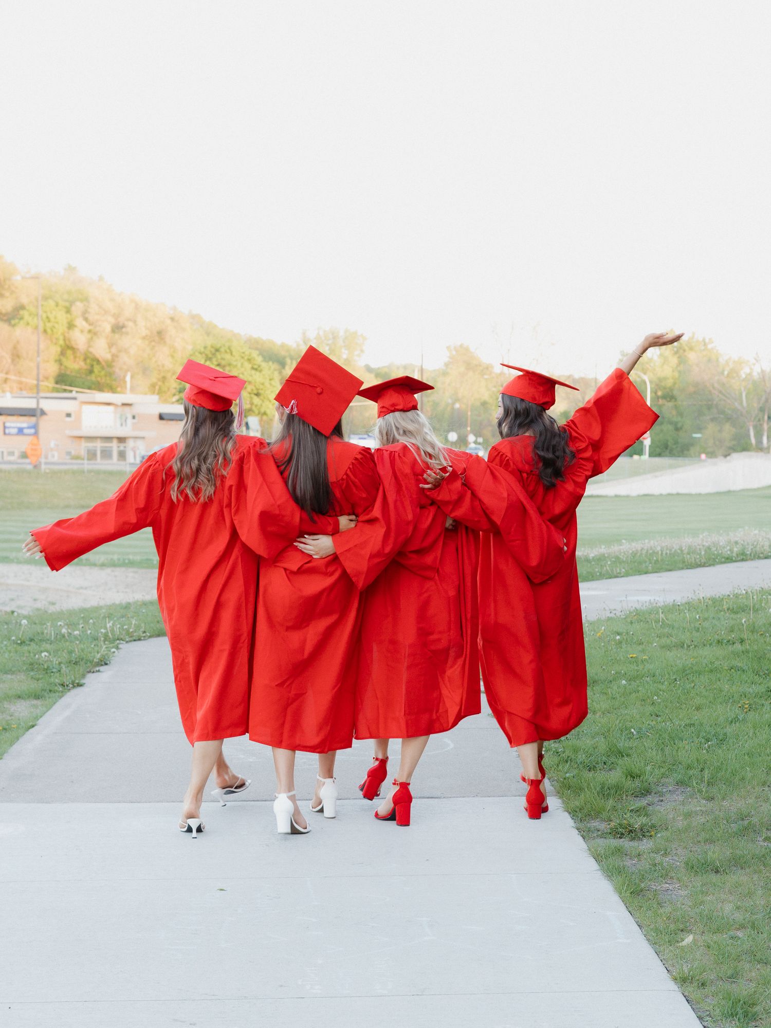 Four female graduates in red caps and gowns walk together on a path, arms linked, celebrating their graduation day.