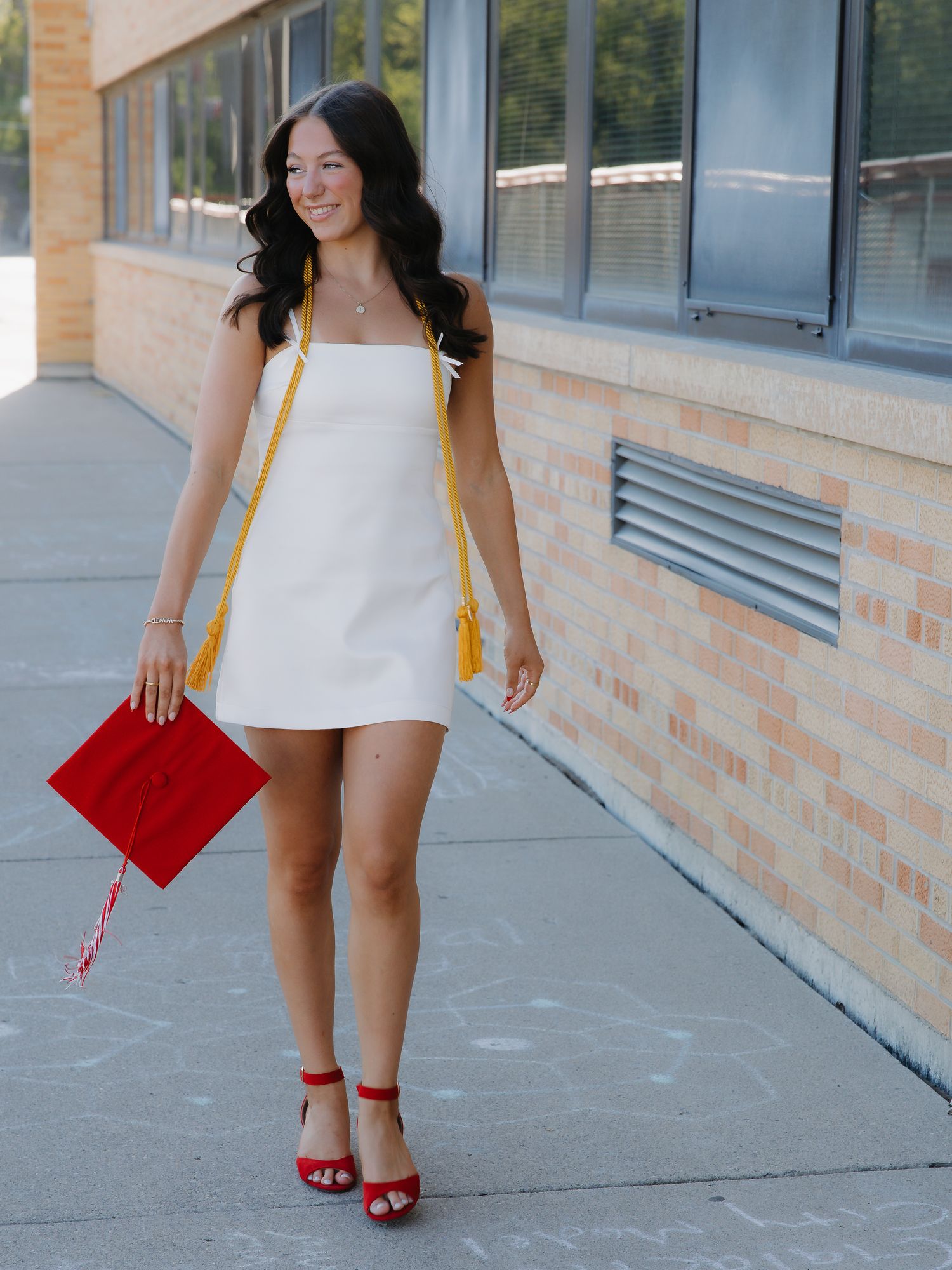 Young woman in white mini dress holding red graduation cap with yellow tassel, posing outdoors near brick building.