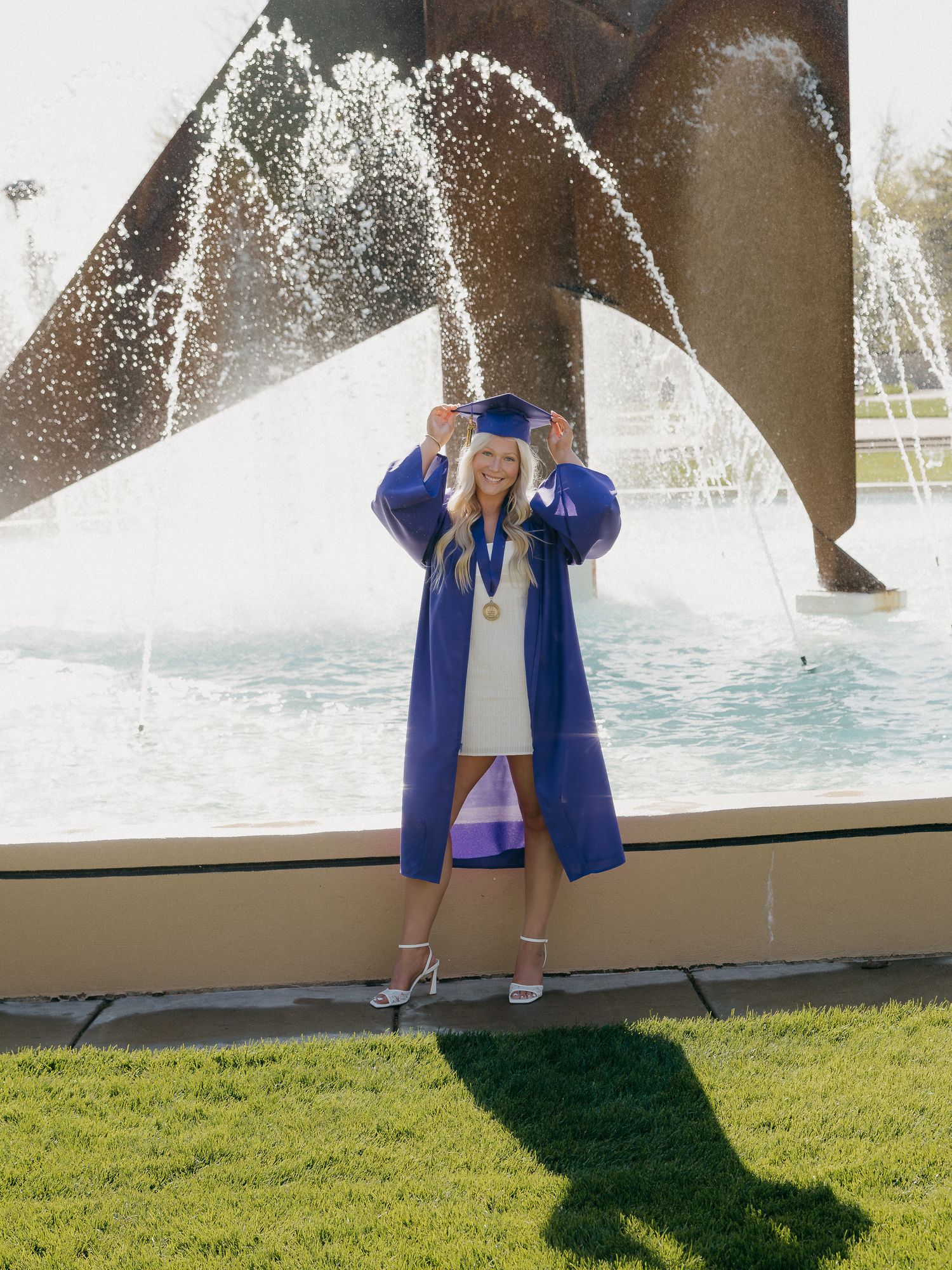 Blonde graduate in purple cap and gown posing in front of a fountain on campus.