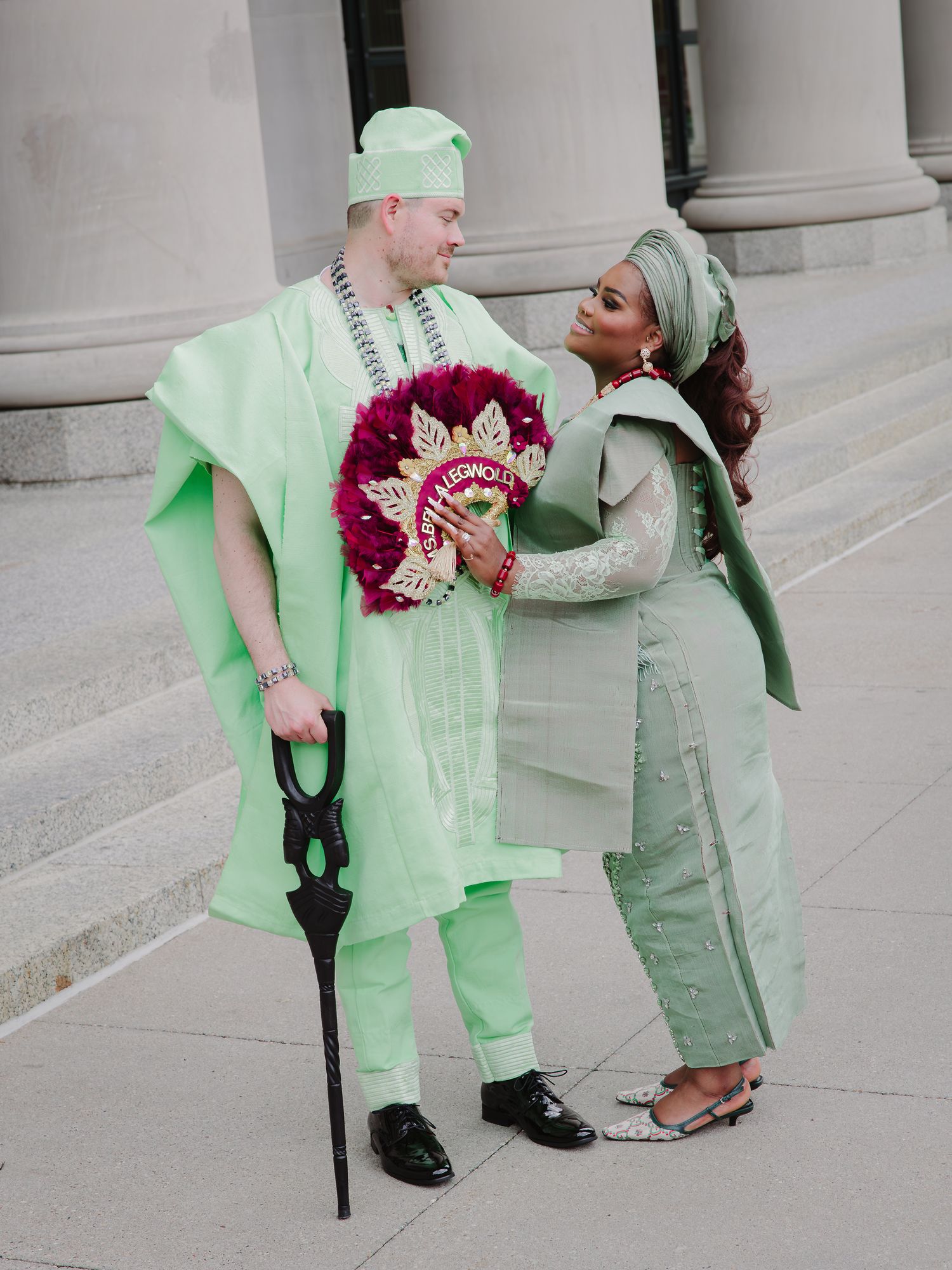 Couple in matching mint green Nigerian traditional wedding attire, holding a decorative bouquet outside stone columns.