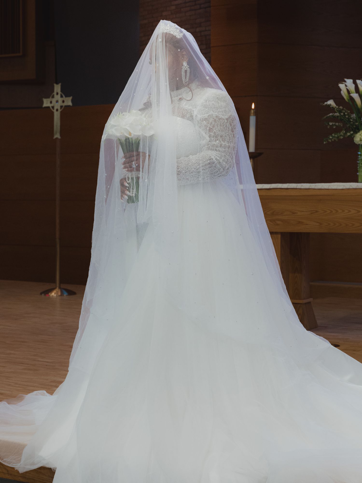Bride in white ball gown and long veil standing at church altar holding bouquet during wedding ceremony.