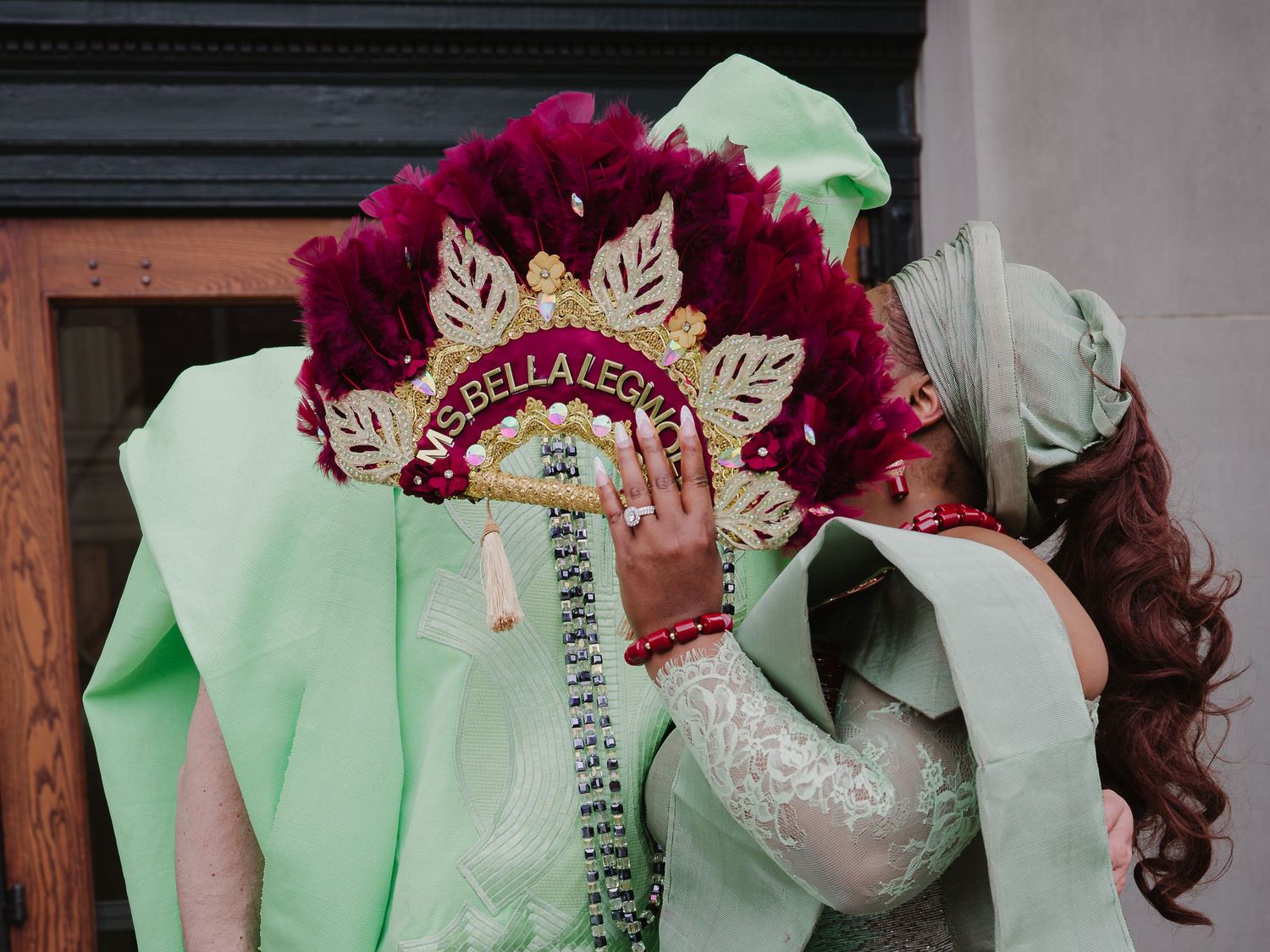 Woman in mint green dress holds ornate crown with red feathers and gold text at a festive celebration.