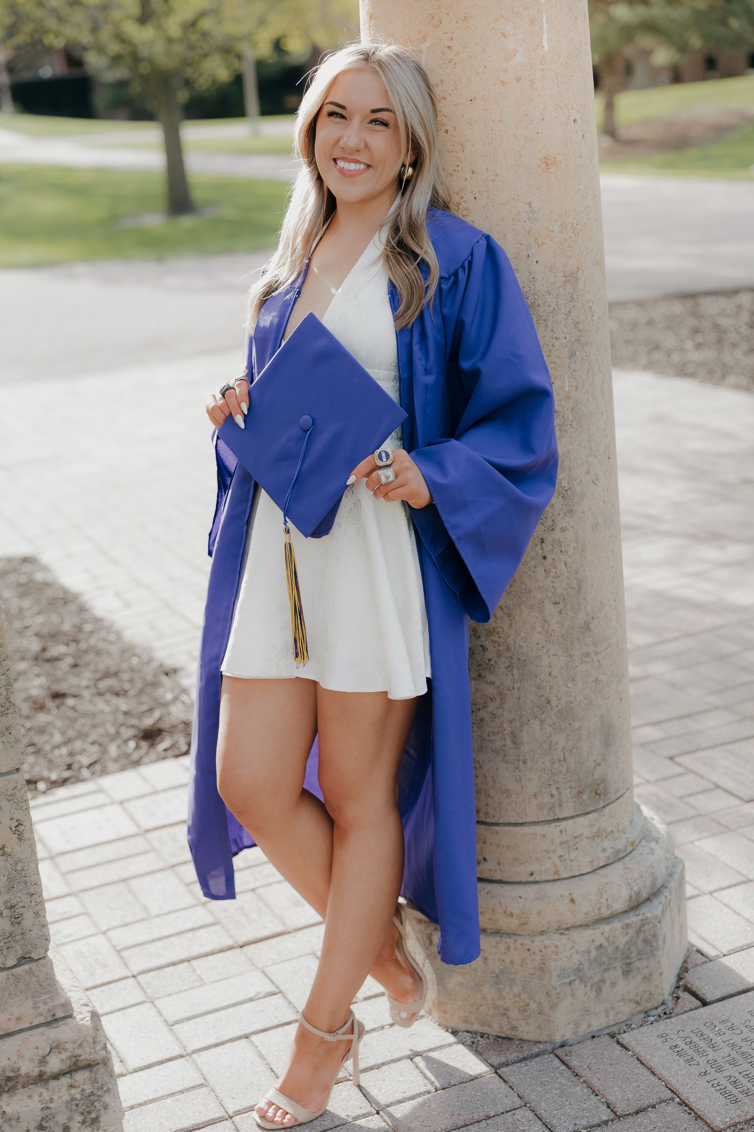 Smiling blonde graduate in blue cap and gown posing against a stone column outdoors in a white dress and heels.