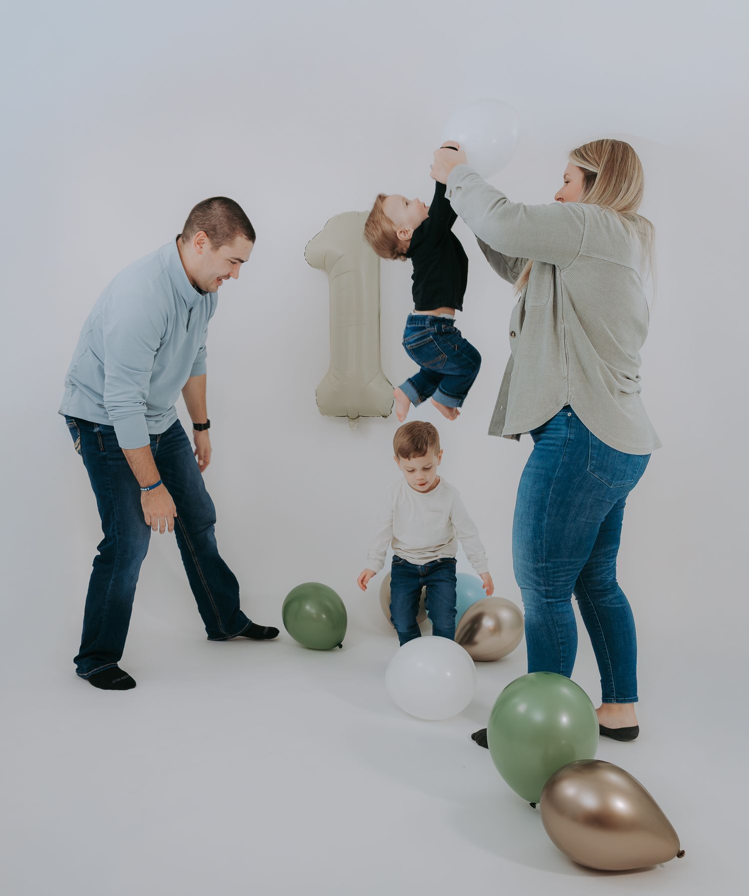 A joyful family of four plays with colorful balloons in a bright white studio, parents lifting a laughing child upside down.