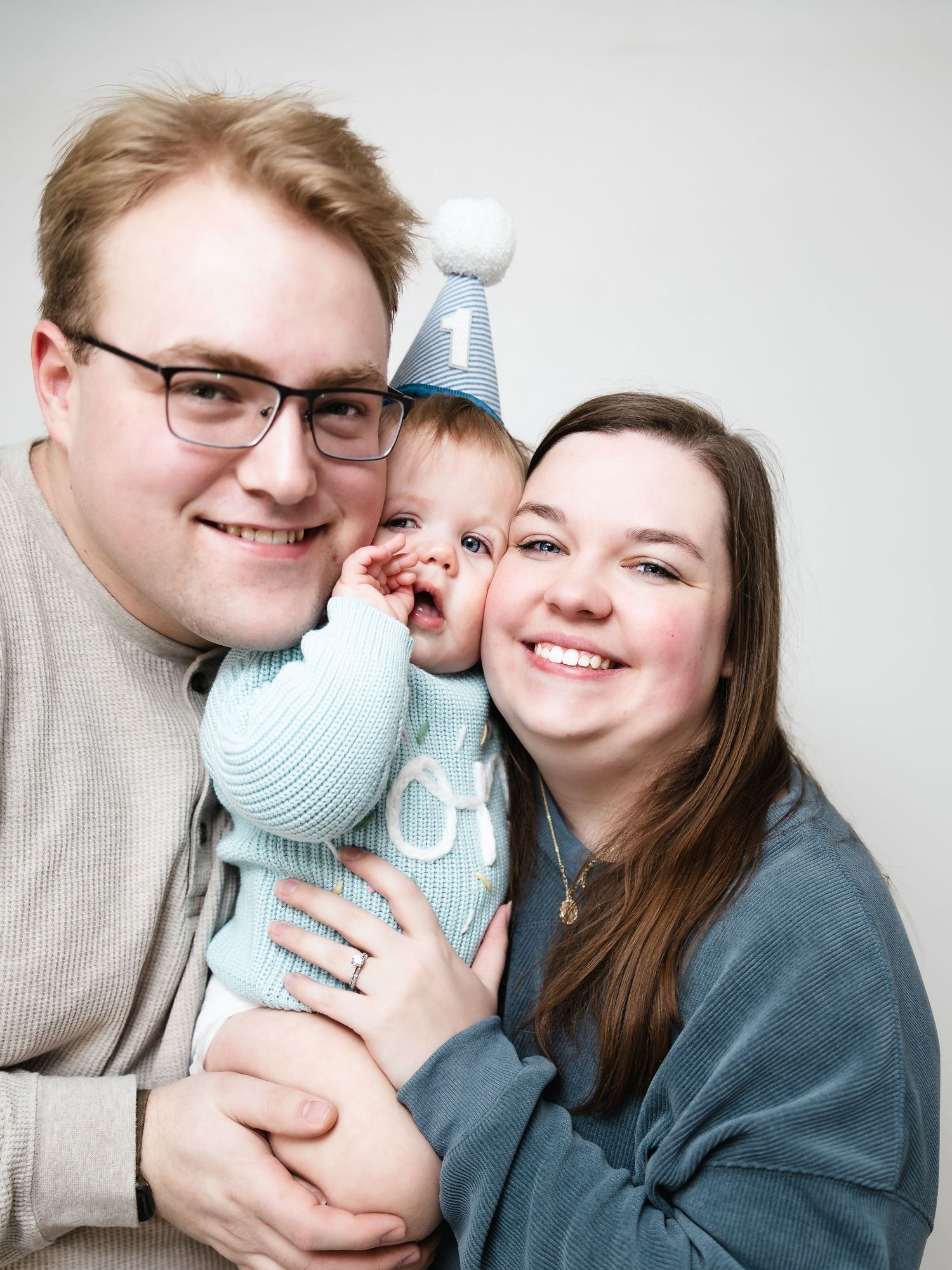 Happy family portrait with parents holding their baby celebrating first birthday with blue party hat.