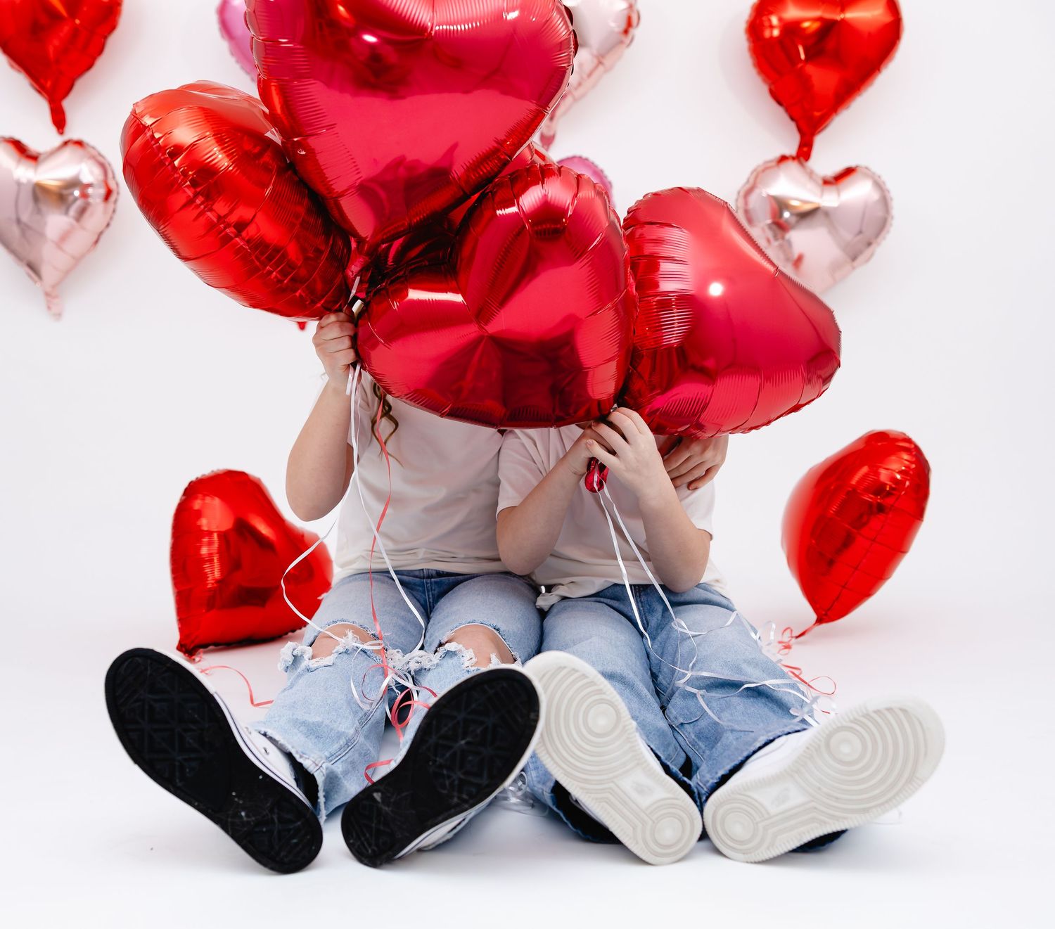 Two children sitting on floor hiding behind red heart-shaped foil balloons on Valentine's Day.