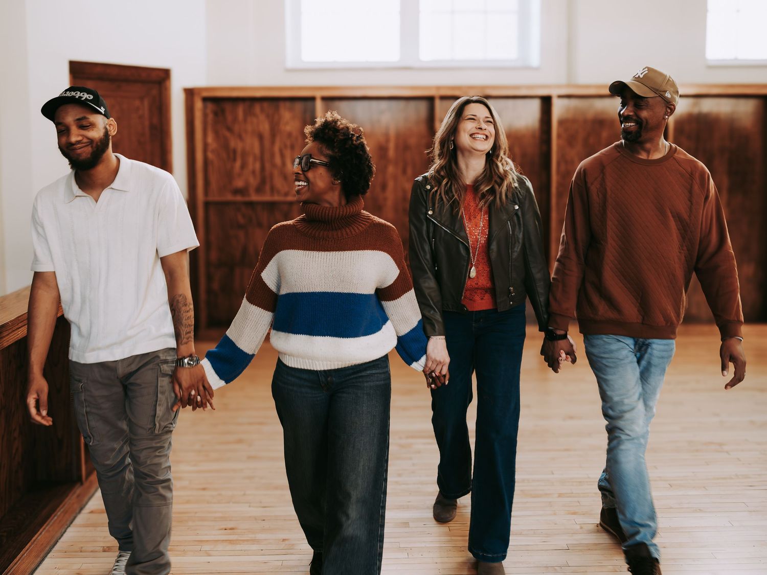 Four diverse friends walk hand in hand laughing together inside a bright wooden room.