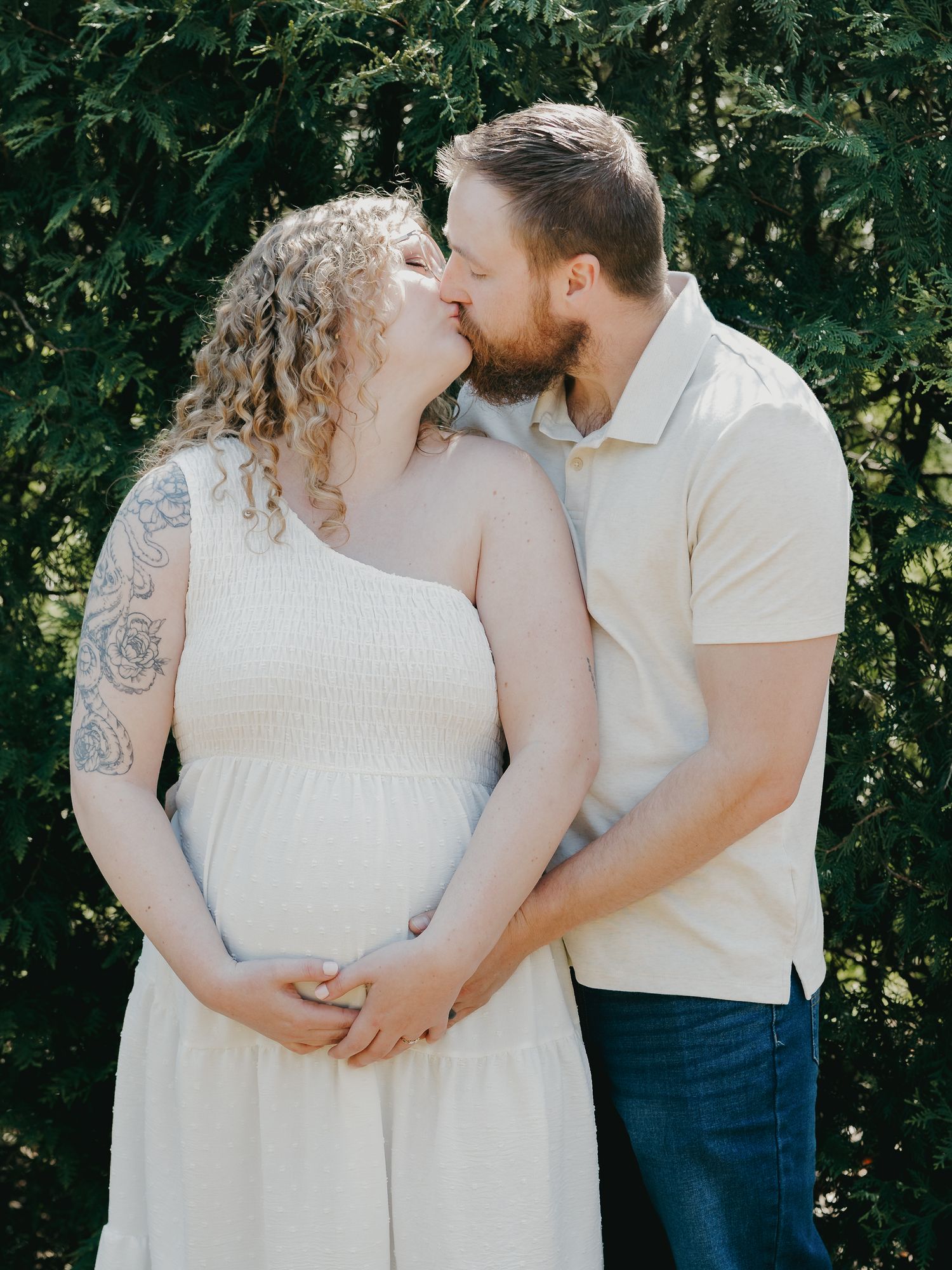Pregnant couple sharing a kiss outdoors, woman in white dress with floral tattoo, man in cream polo and jeans, green trees behind.