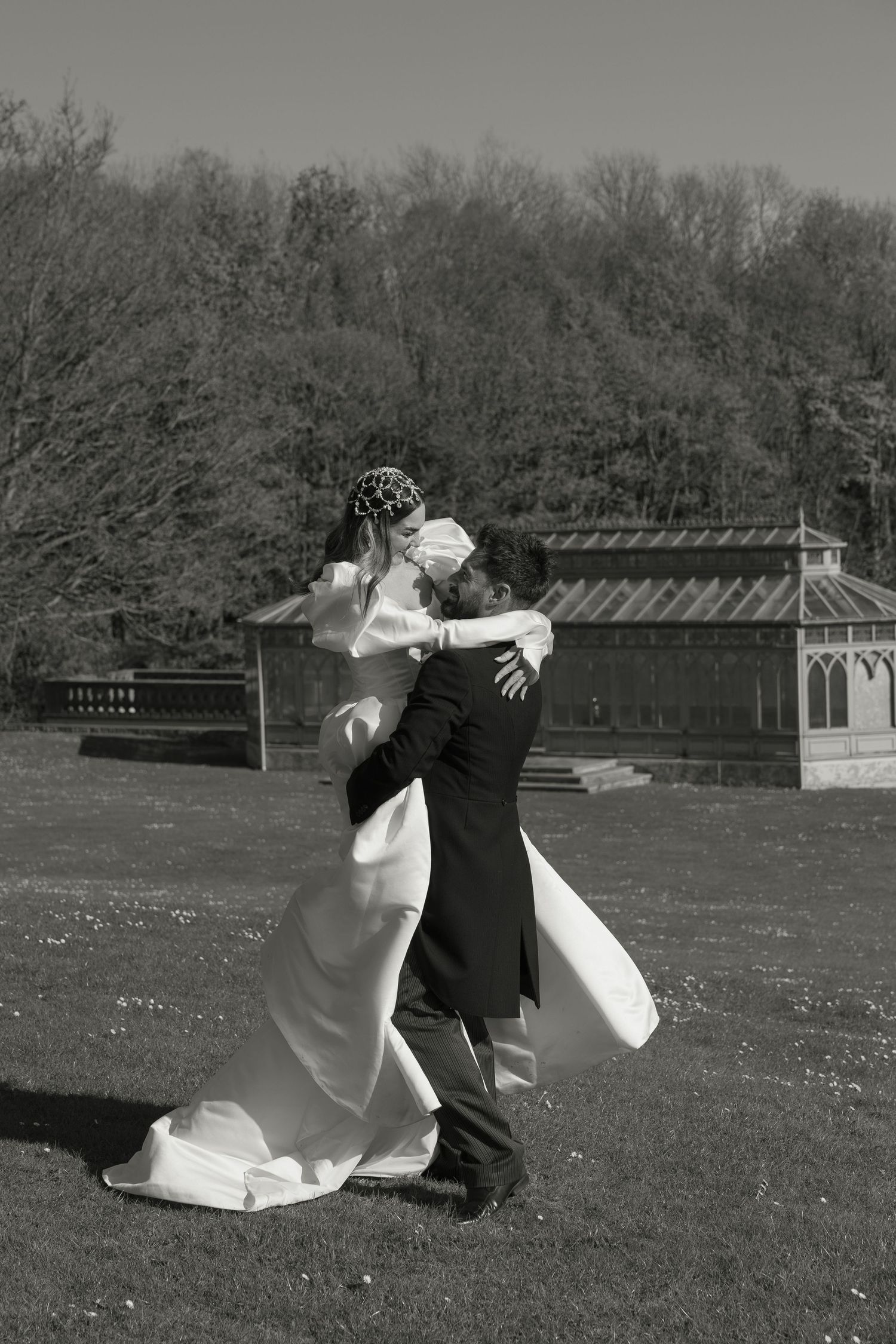 Black and white wedding photo of bride and groom embracing outdoors on sunny day in front of garden pavilion.