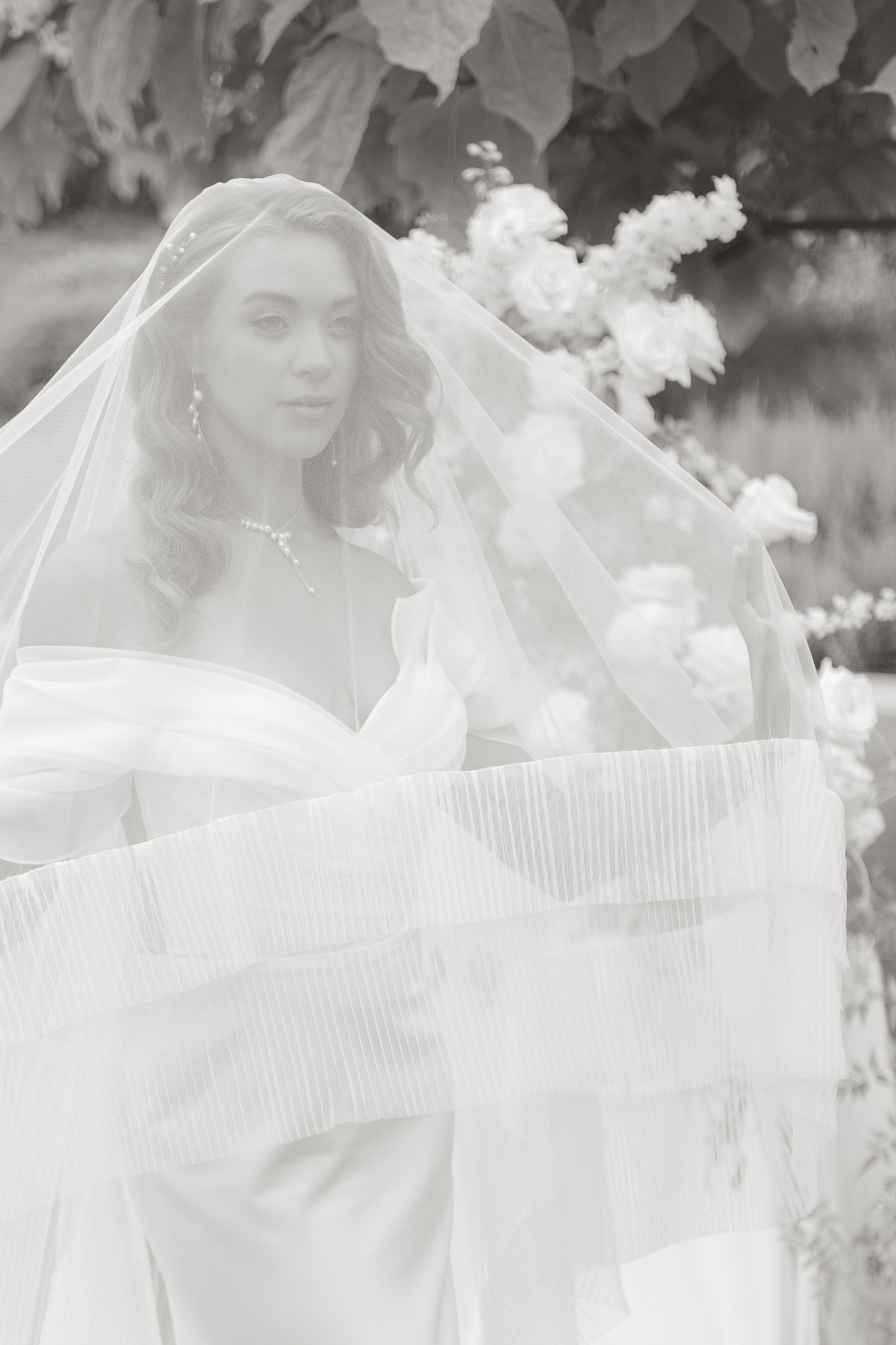 Bride in off-shoulder gown draped with flowing veil among white blooms, black and white wedding portrait.