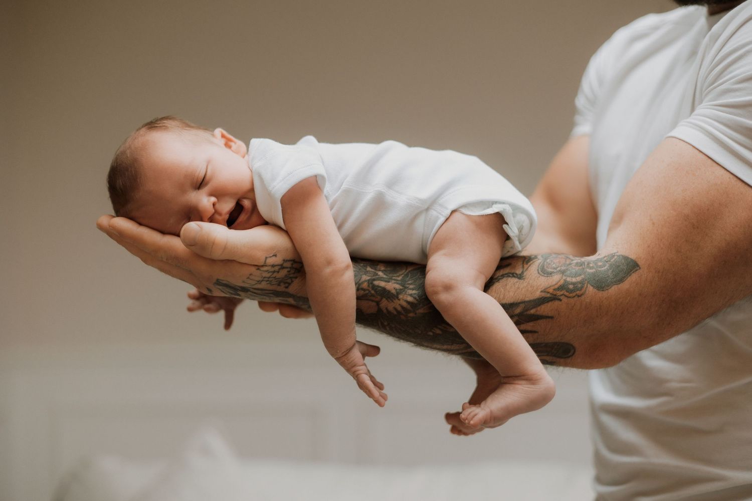 Tattooed father carefully holding sleeping newborn baby in white onesie with both hands.
