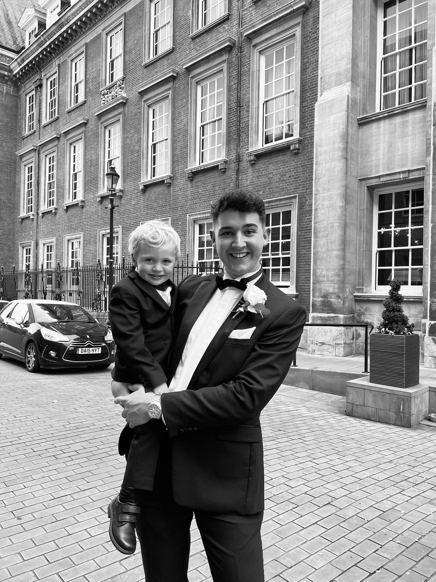 Young man in tuxedo holding a small boy in formal wear outside an elegant brick building.