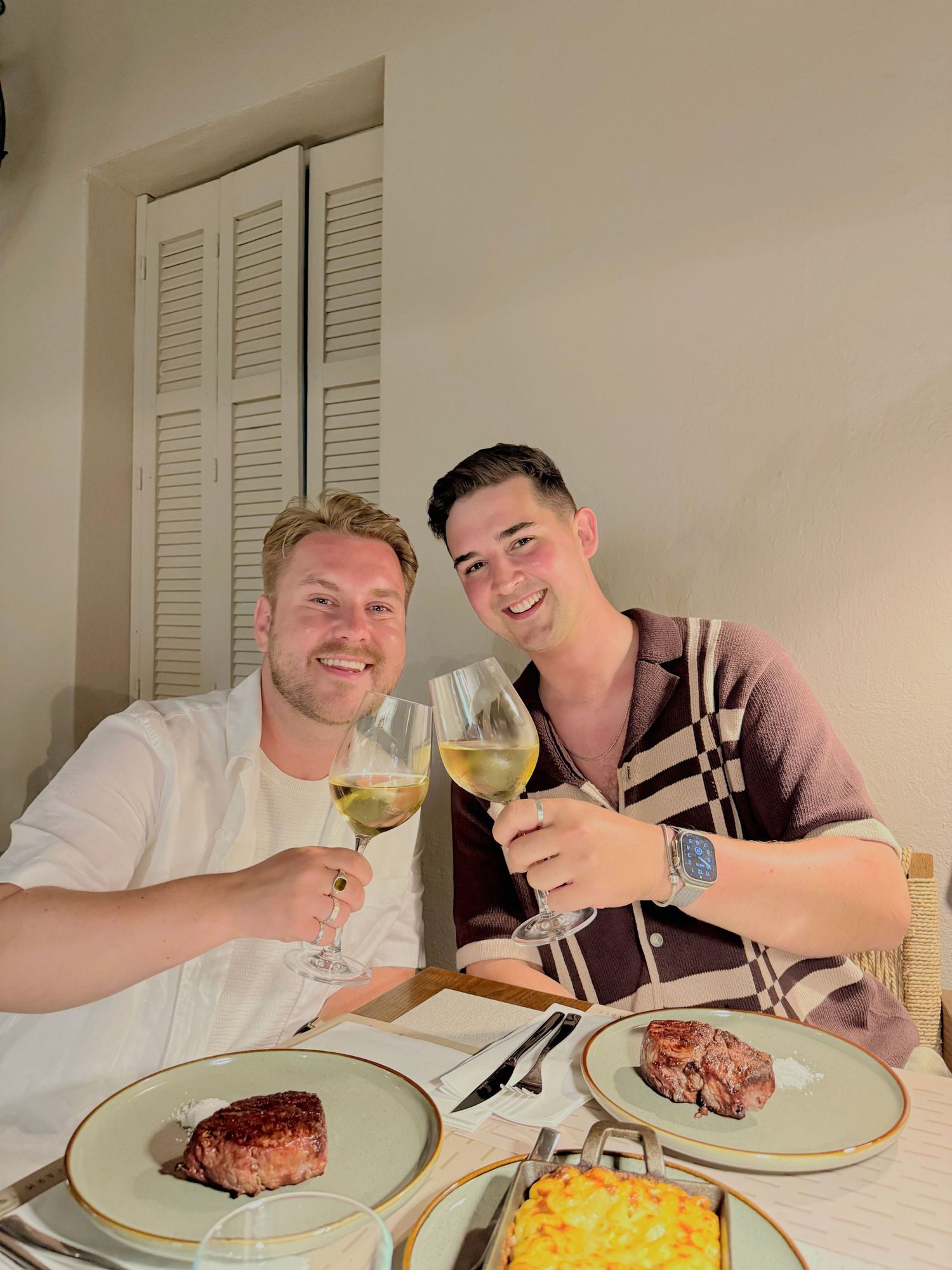Two men toasting with white wine glasses over steak dinner at a restaurant table.