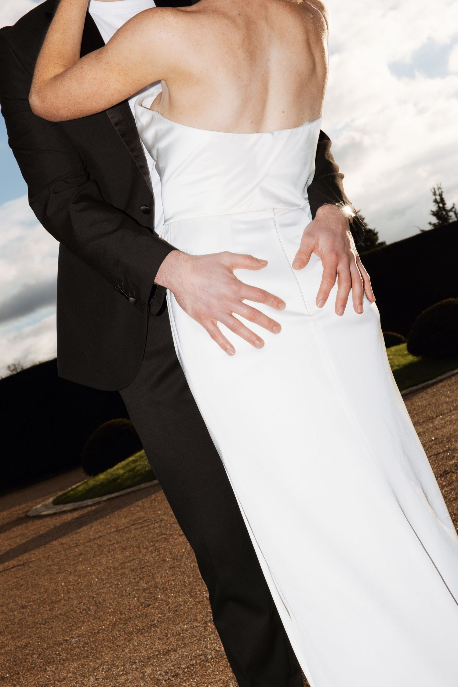 Close-up of wedding couple embracing, groom's hands on bride's white strapless gown waist outdoors.