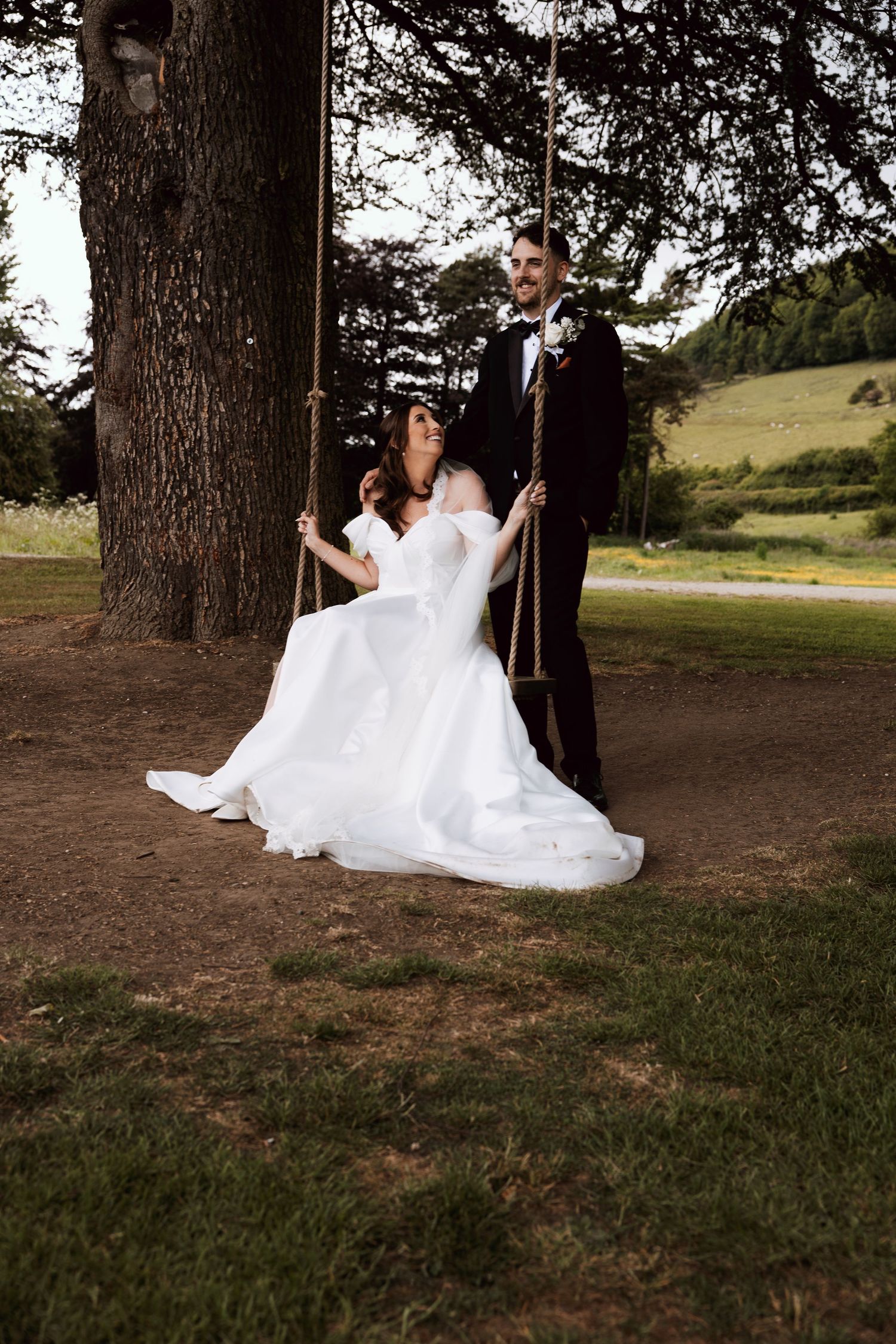 Bride in white off-shoulder gown sits on rope swing as groom in black tuxedo stands beside her outdoors.