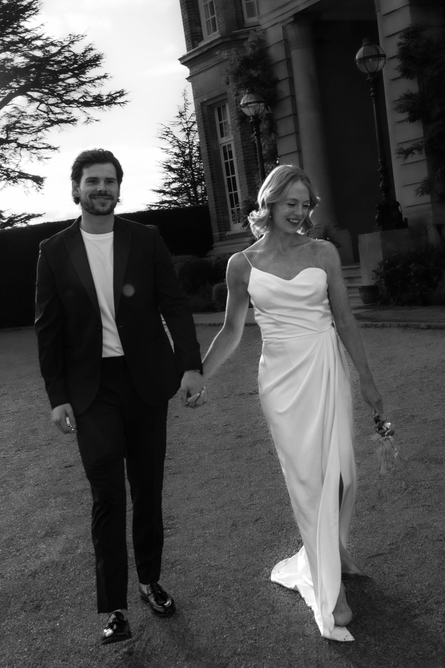 Black and white photo of smiling bride and groom holding hands walking outside a grand manor house.