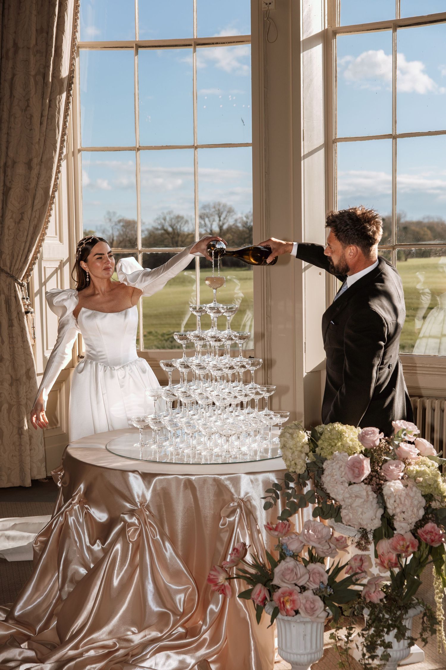 Bride and groom pouring champagne tower at elegant wedding venue with large windows and floral arrangements.