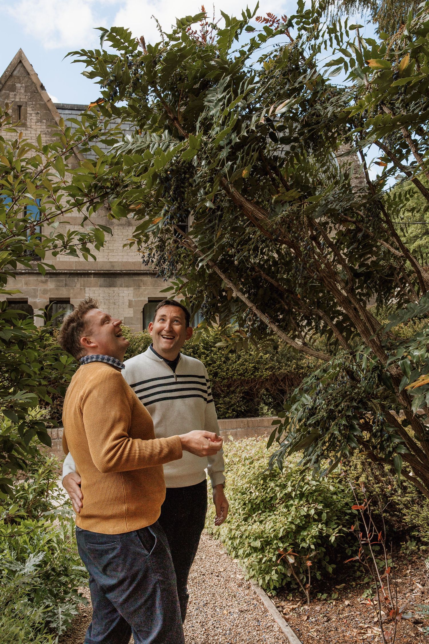 Two men laughing together in a lush garden beside a stone building, one in orange sweater, one in striped vest.