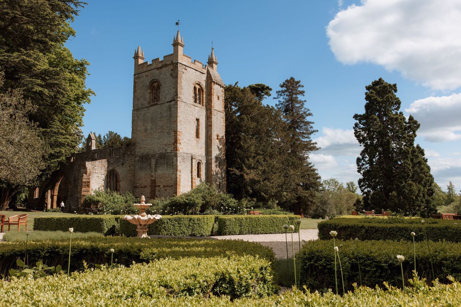 Historic stone castle tower with manicured hedges, fountain, and tall trees under a blue sky.
