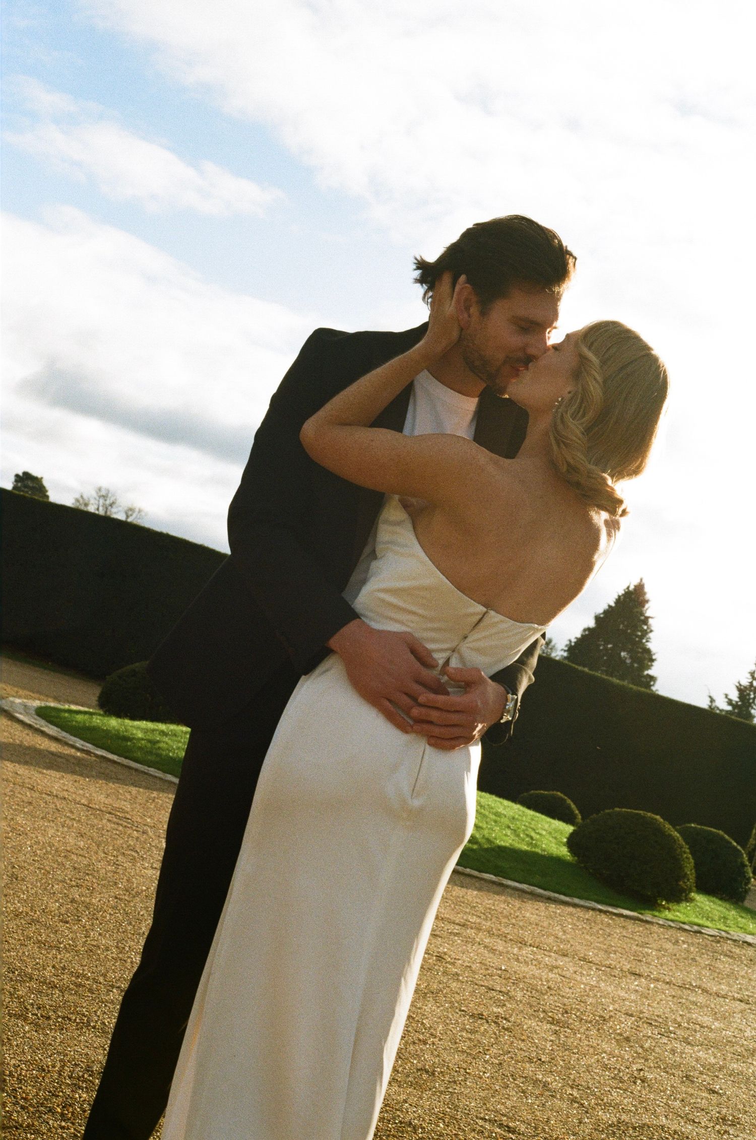 Bride and groom sharing a romantic kiss at golden hour, bride in sleek backless white gown outdoors.