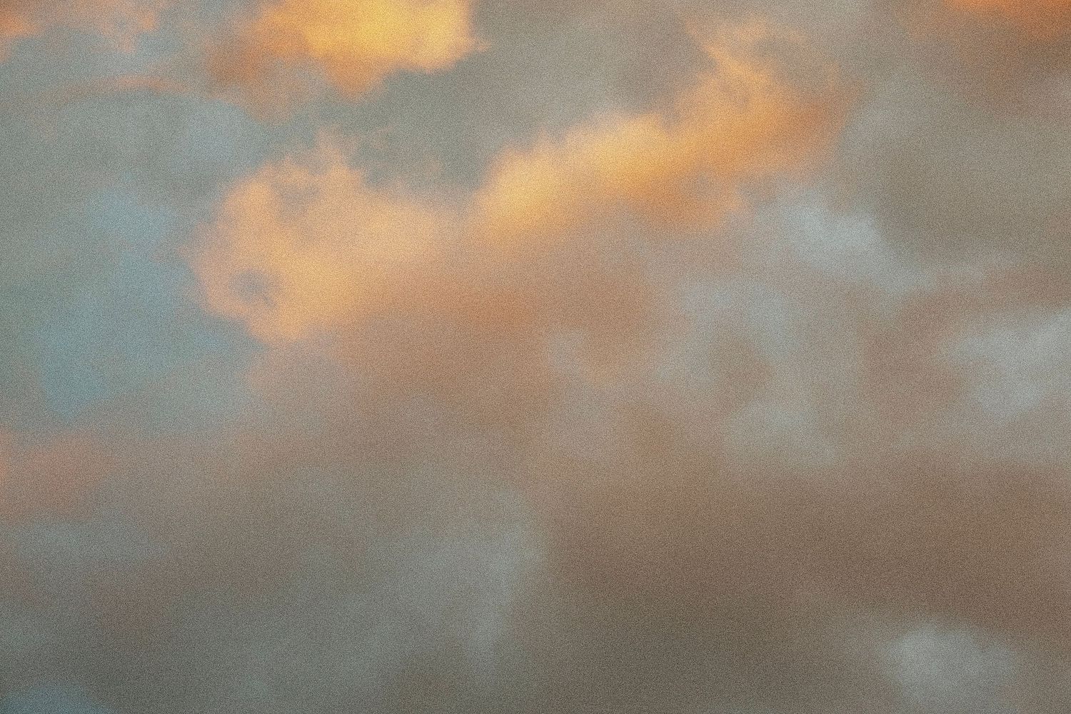 Soft pastel clouds illuminated by warm golden sunset light fill a dreamy sky with shades of orange, pink, and gray.
