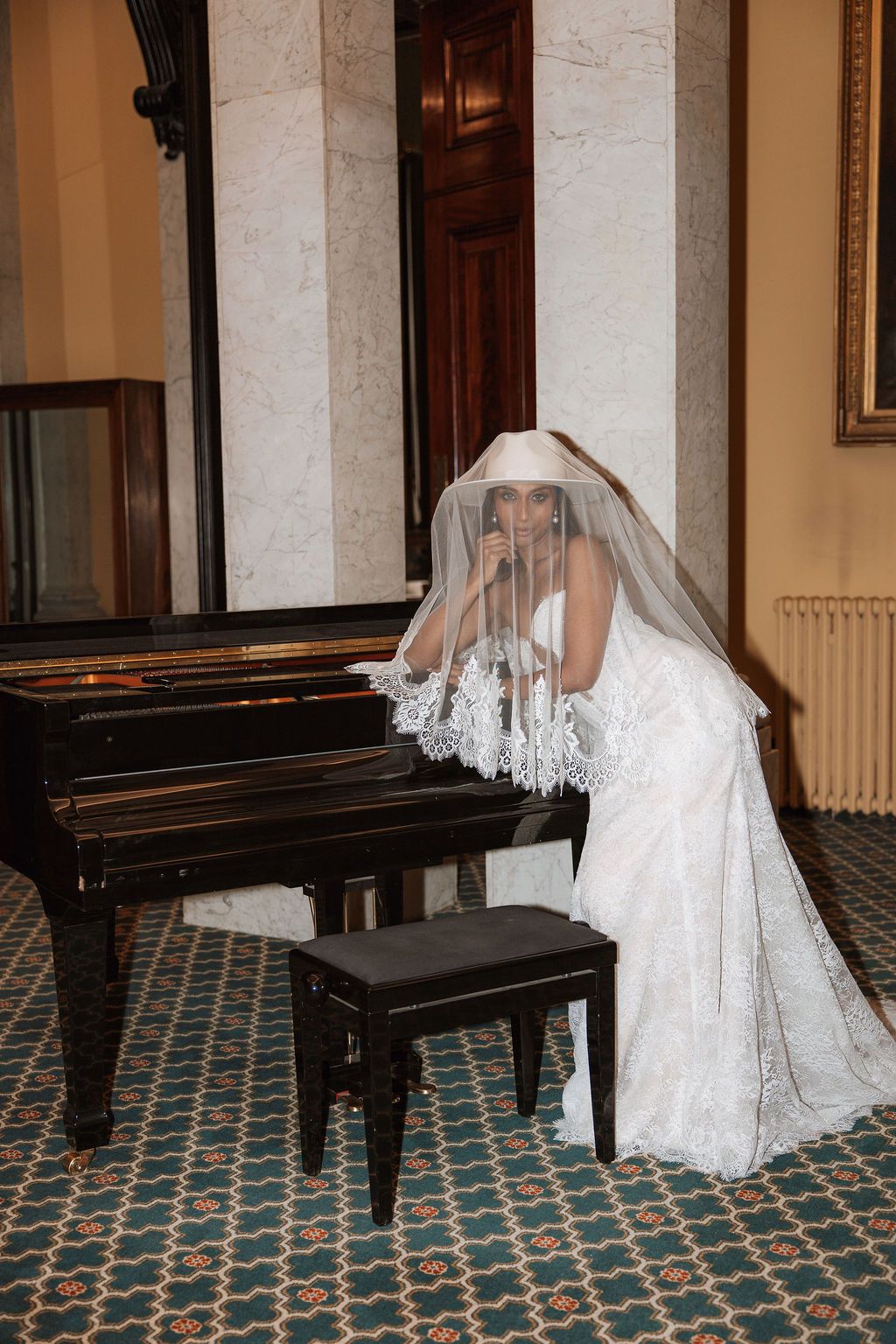Bride in white lace wedding dress and veil poses elegantly beside a grand piano in an ornate room.