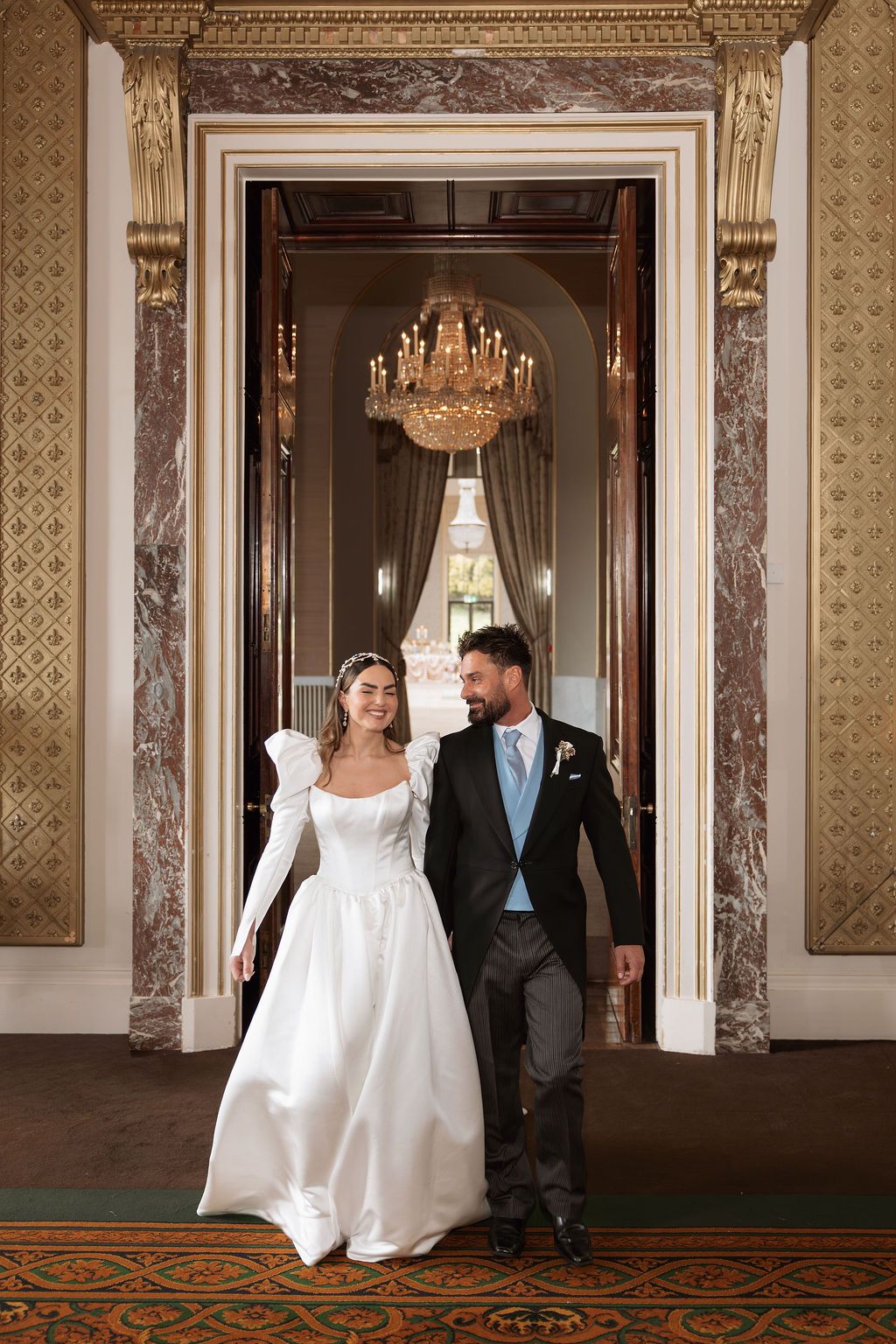 Elegant bride and groom walking through ornate marble doorway in grand ballroom with crystal chandelier.