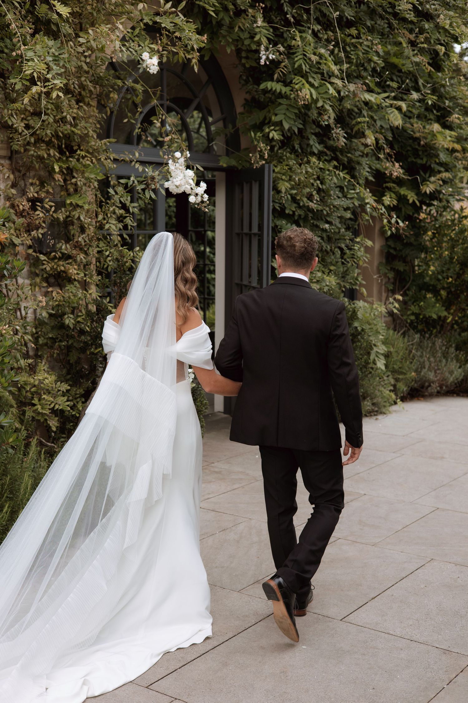 Bride and groom walking hand in hand through lush garden archway, bride in off-shoulder gown with long veil.