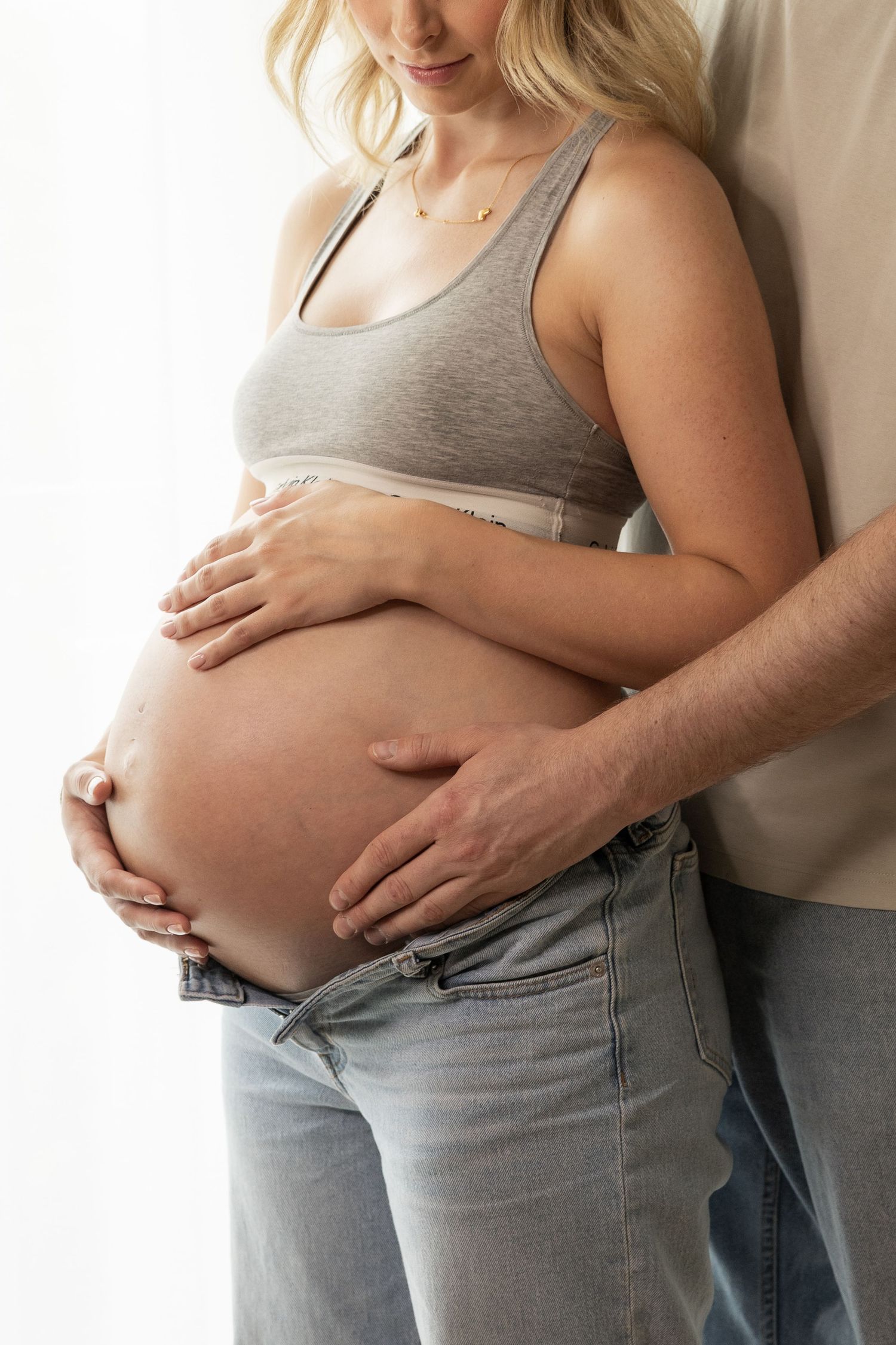 Close-up of pregnant womans bare belly with partners hands gently cradling bump near window.